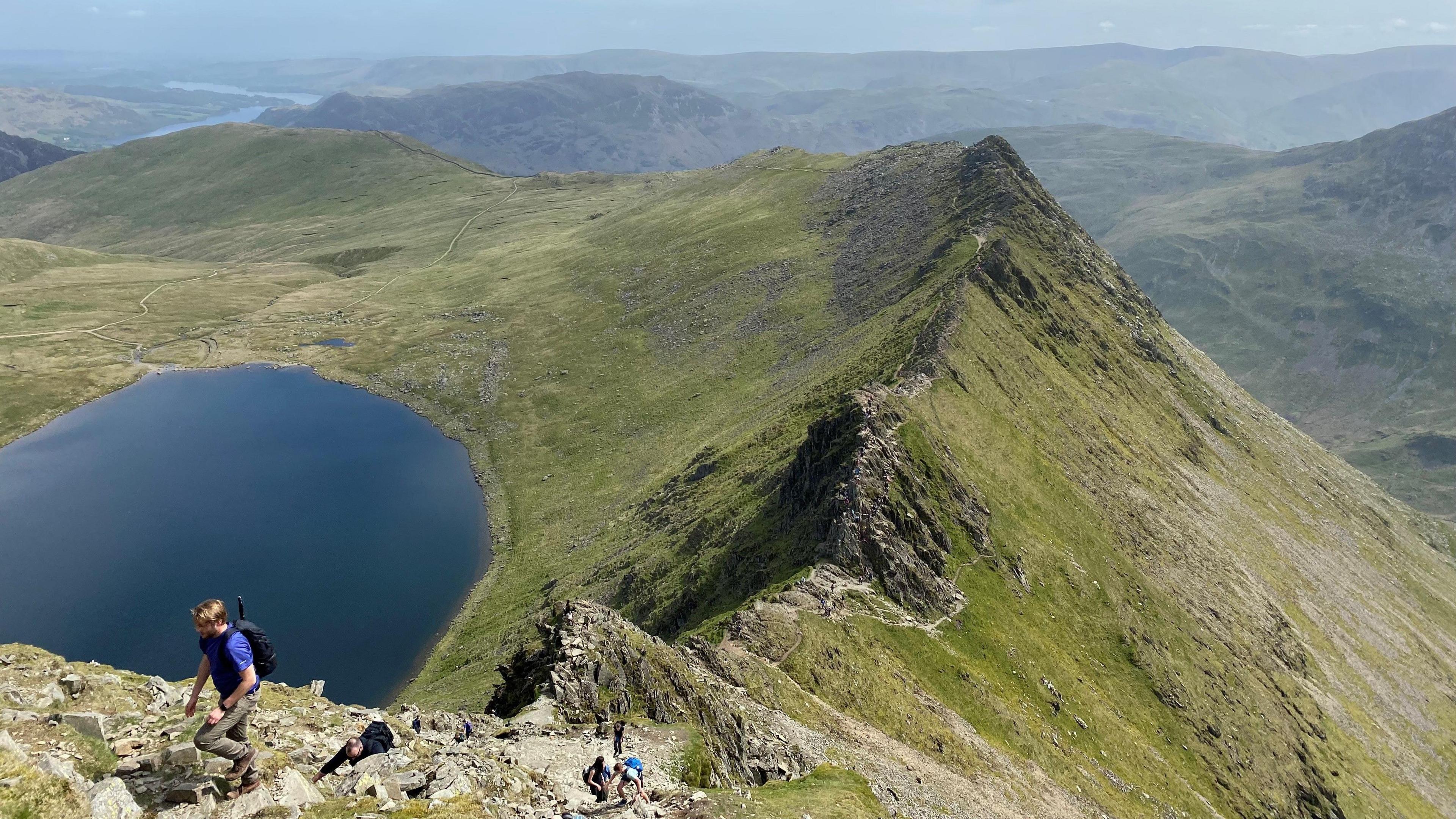 A walker in a blue shirt nears the top of a steep rocky hill. Beyond him is a craggy line of rocks with steep sides either side sloping down. The slope on the left ends in a large blue tarn. There are other hills beyond.