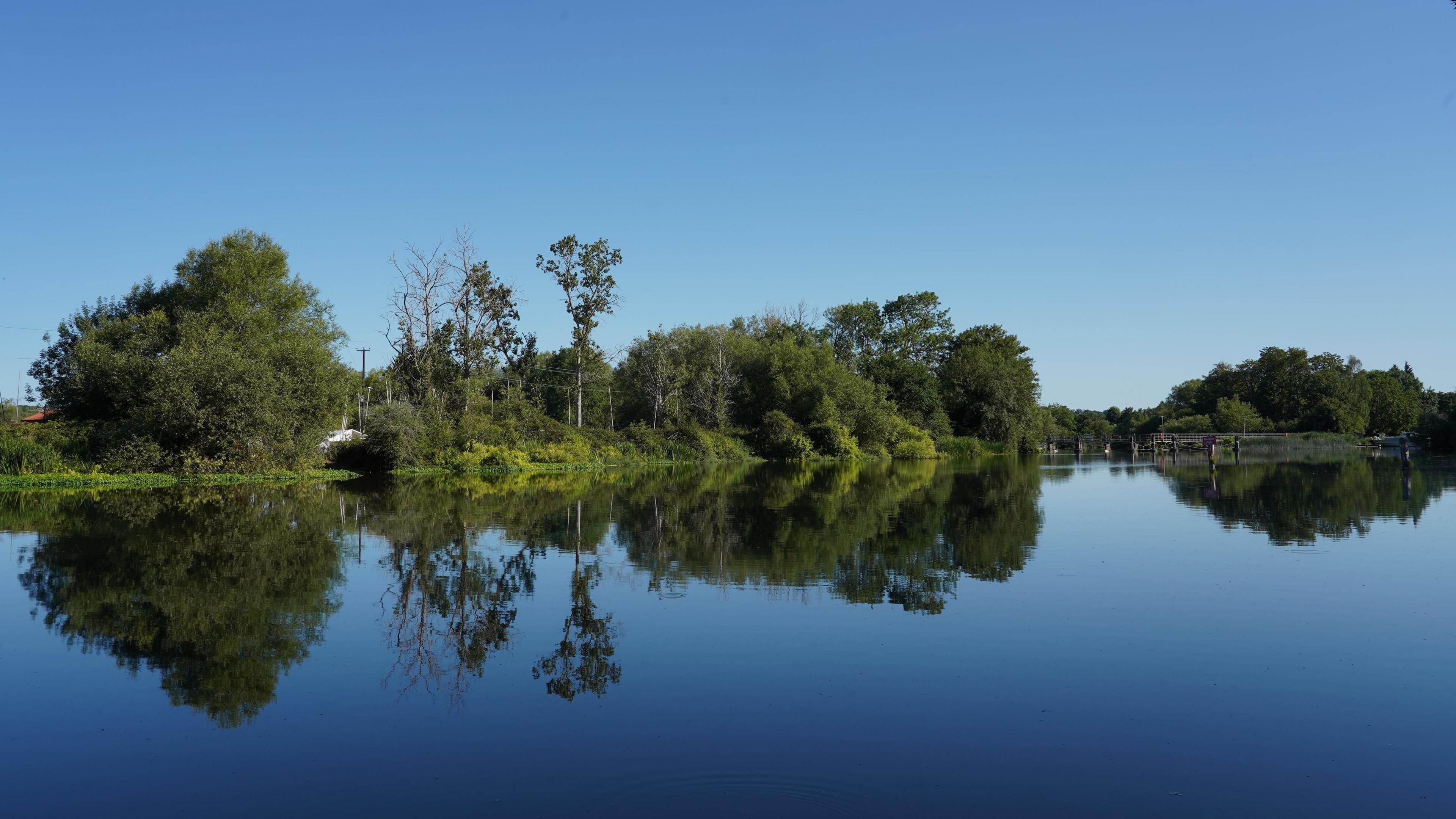 A copse of trees runs through the middle of this shot with a weir just visible in the background. The cloudless blue skies are reflected in the mirror like river water as is the copse giving a perfect symmetry to the image.