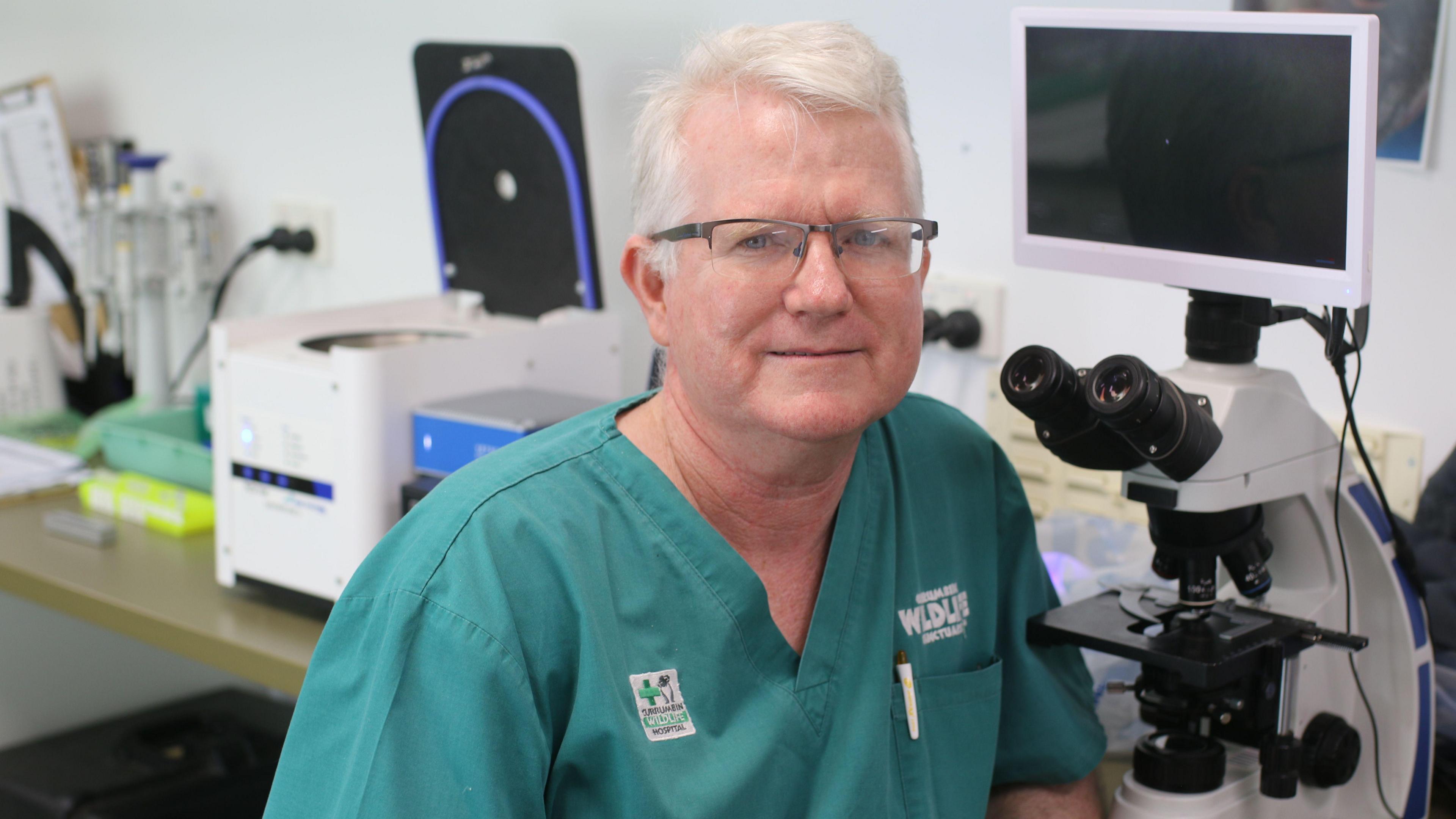 A man with glasses and white hair in green scrubs looks at the camera and smiles. He is sitting in front of a desk with medical equipment including a microscope on it.