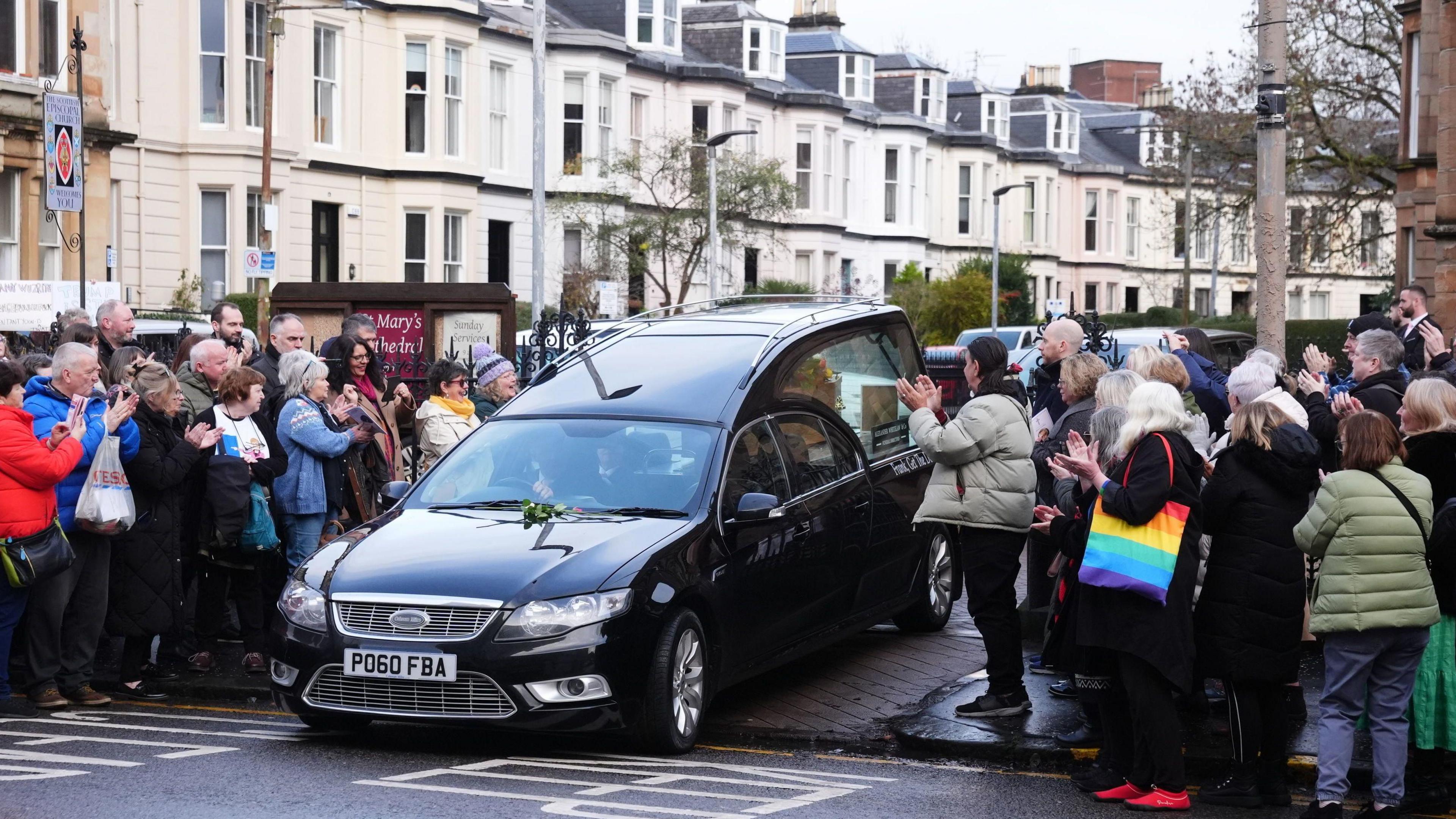 A black hearse in the middle of a road with  a small crowd of people wearing warm looking jackets and carrying bags clapping their hands.