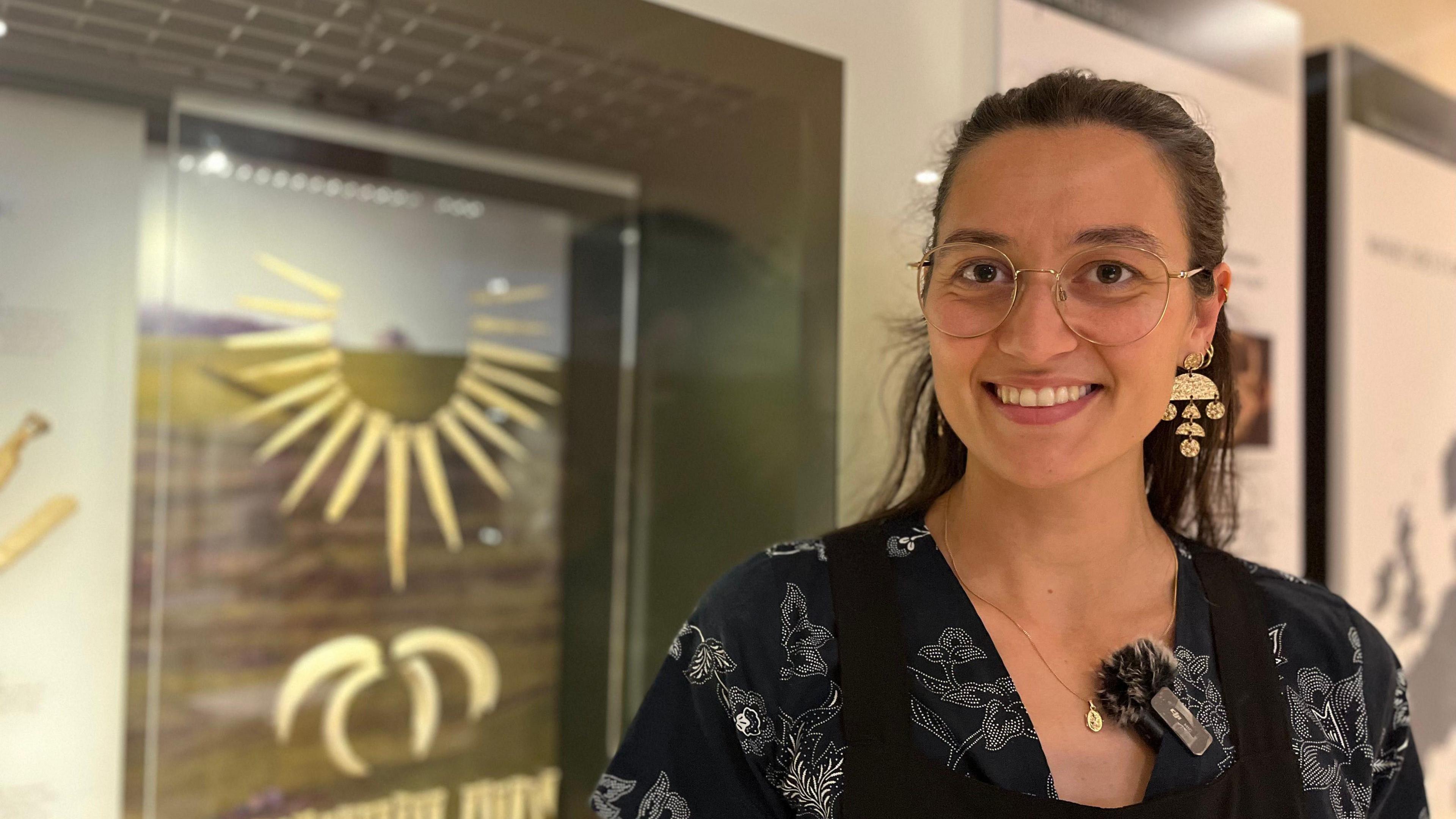 Amber smiles at the camera. She stands in front of a blurred cabinet with items in.
She wears glasses and black floral top.