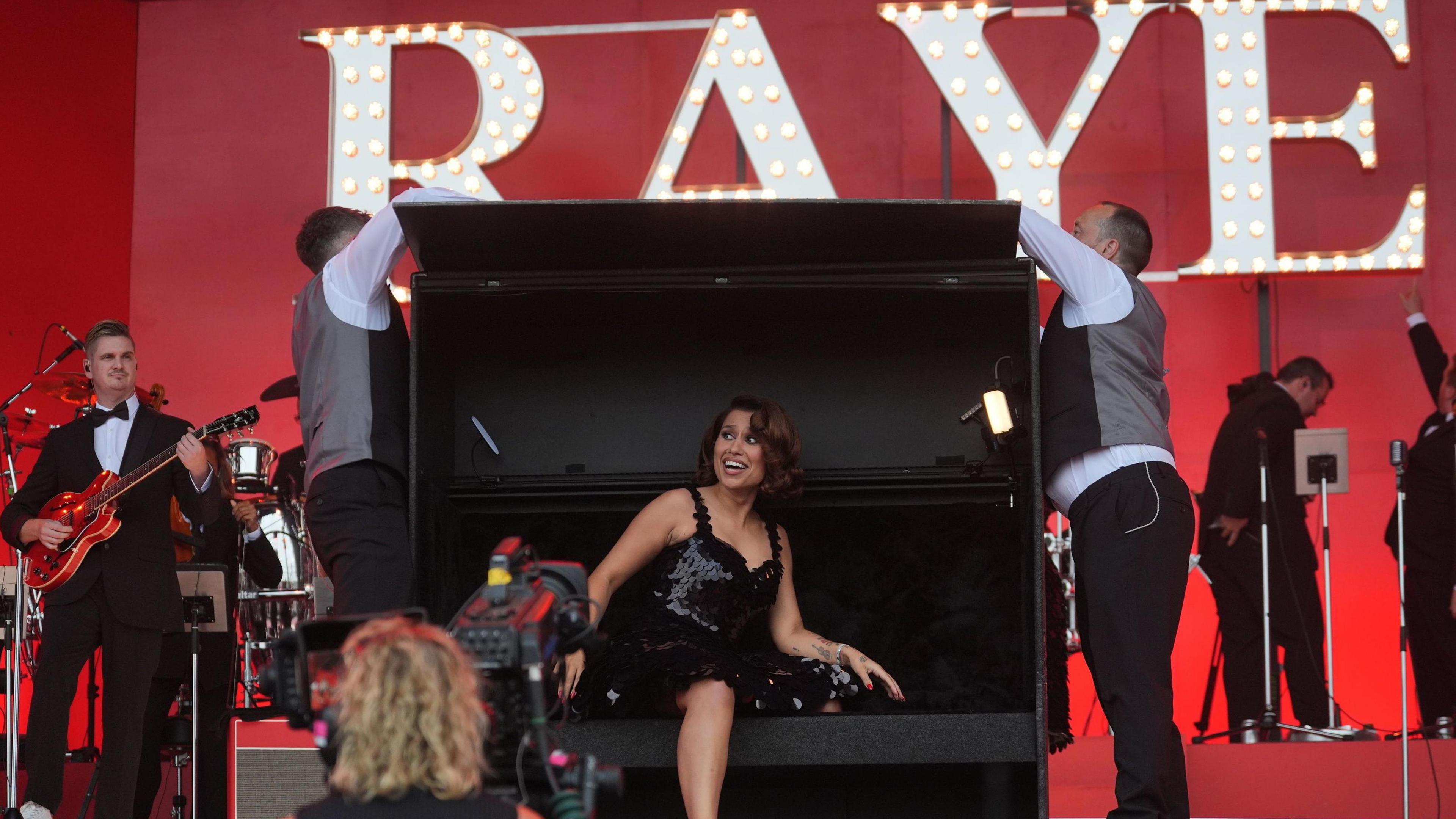 Singer Raye is sat inside a box which has just been opened by two men in smart attire. She is wearing a sequined black dress, with thin straps. Her hair is curled in a 1950s style. The backdrop is all red with 'RAYE' written in bright lights.