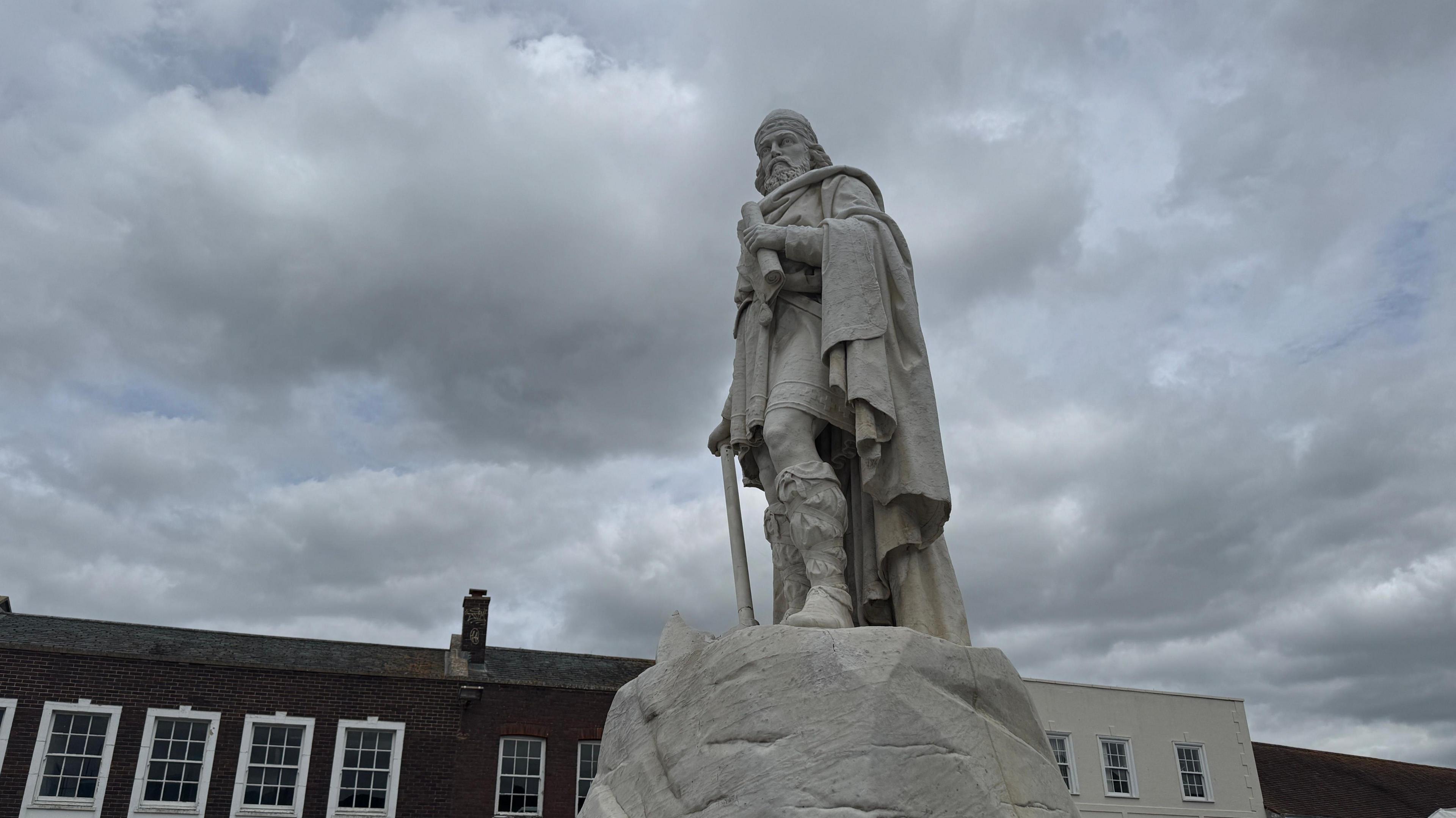 The statue of King Alfred is pictured on a plinth and taken from the ground nearby. A roof is visible in the background the skies are grey and overcast.