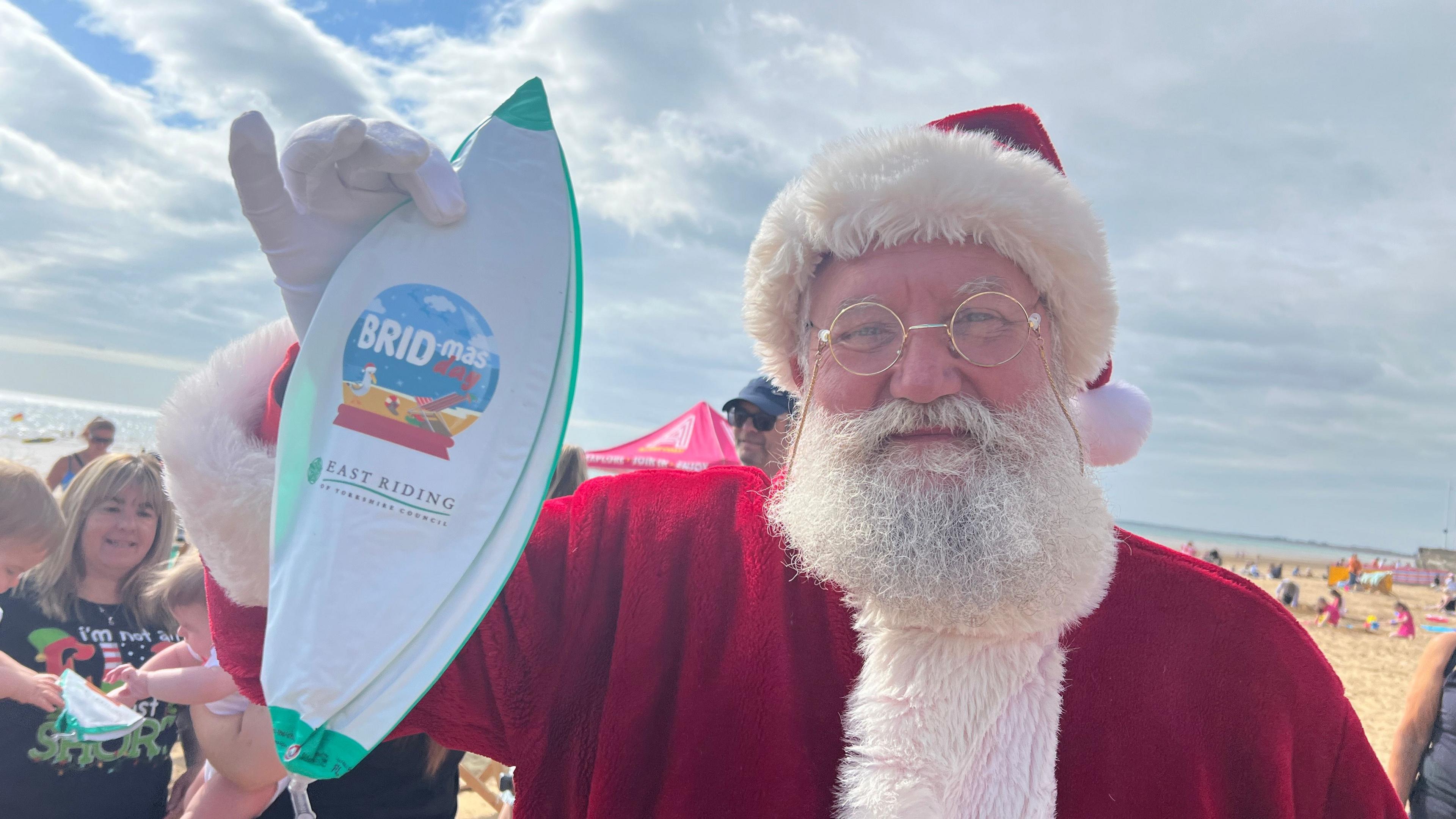 A man dressed as Santa Clause holds up some Bridlington themed promotional materials on the beach.