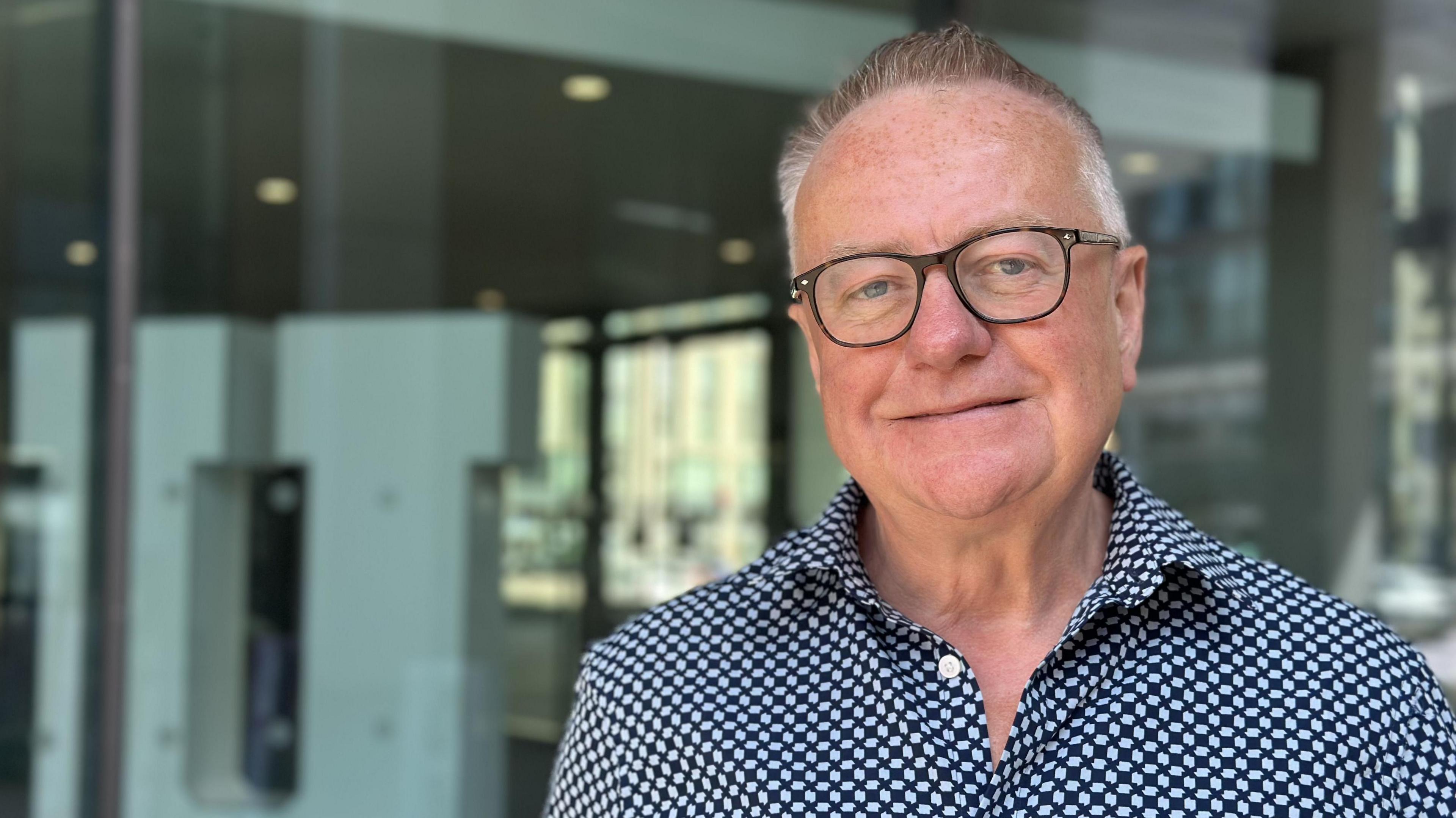 Professor Paddy Gray is wearing glasses and a blue checked shirt. He has short grey hair and he is smiling at the camera. Professor Gray is standing in front of a window at Ulster University's campus in Belfast.