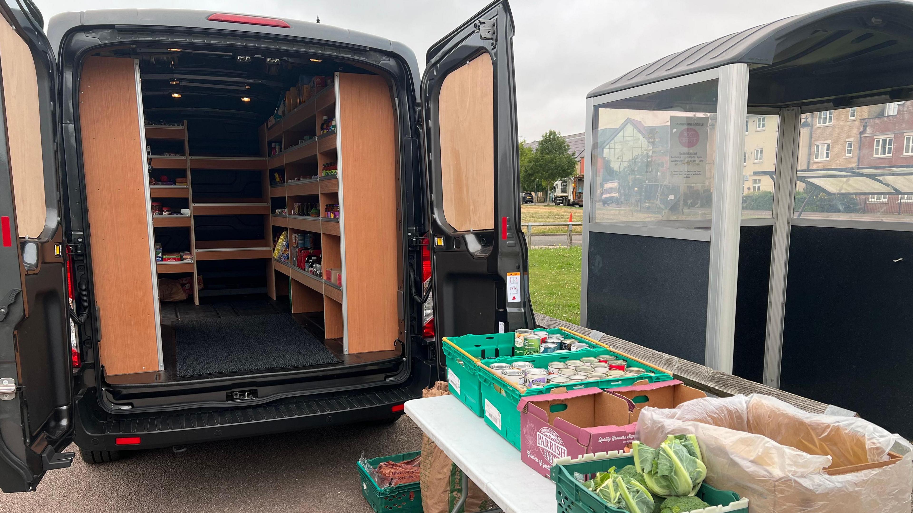 The back of a van with doors open showing shelves full of goods and behind it a table with crates of tinned and fresh vegetables