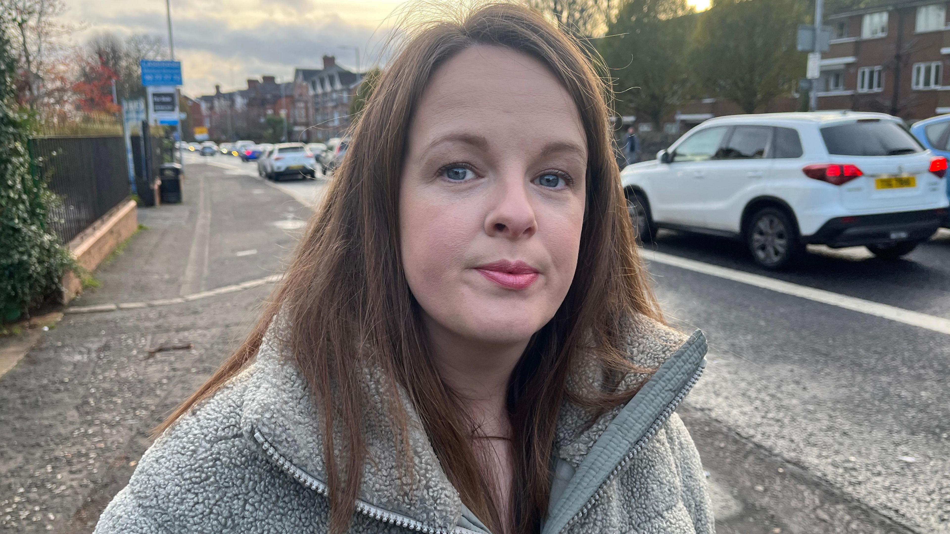 A woman with brown hair, she is wearing a green jacket. She is standing in a street. 