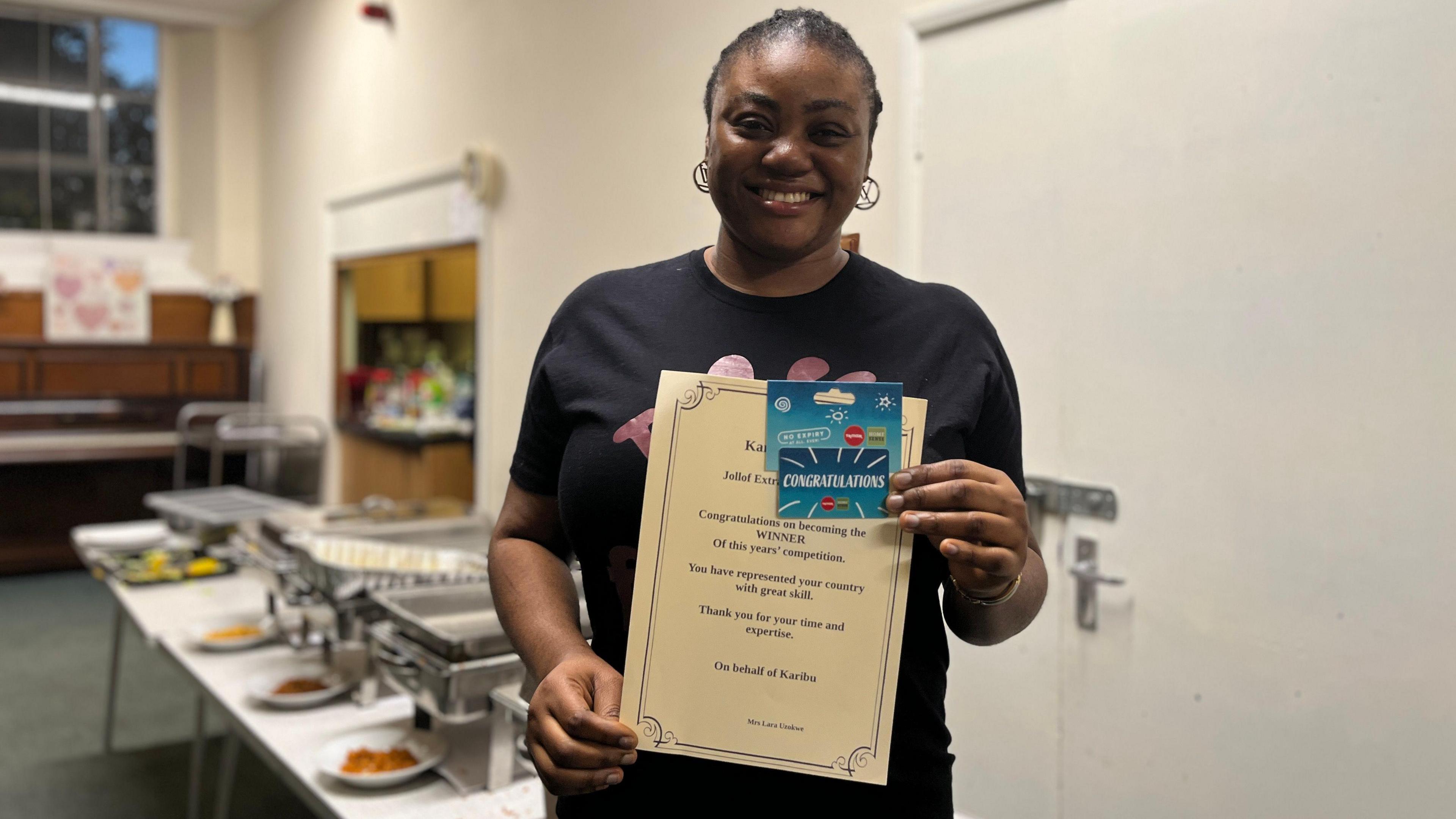 Omolara Agboola stands holding a winner's certificate in front of tables of food.