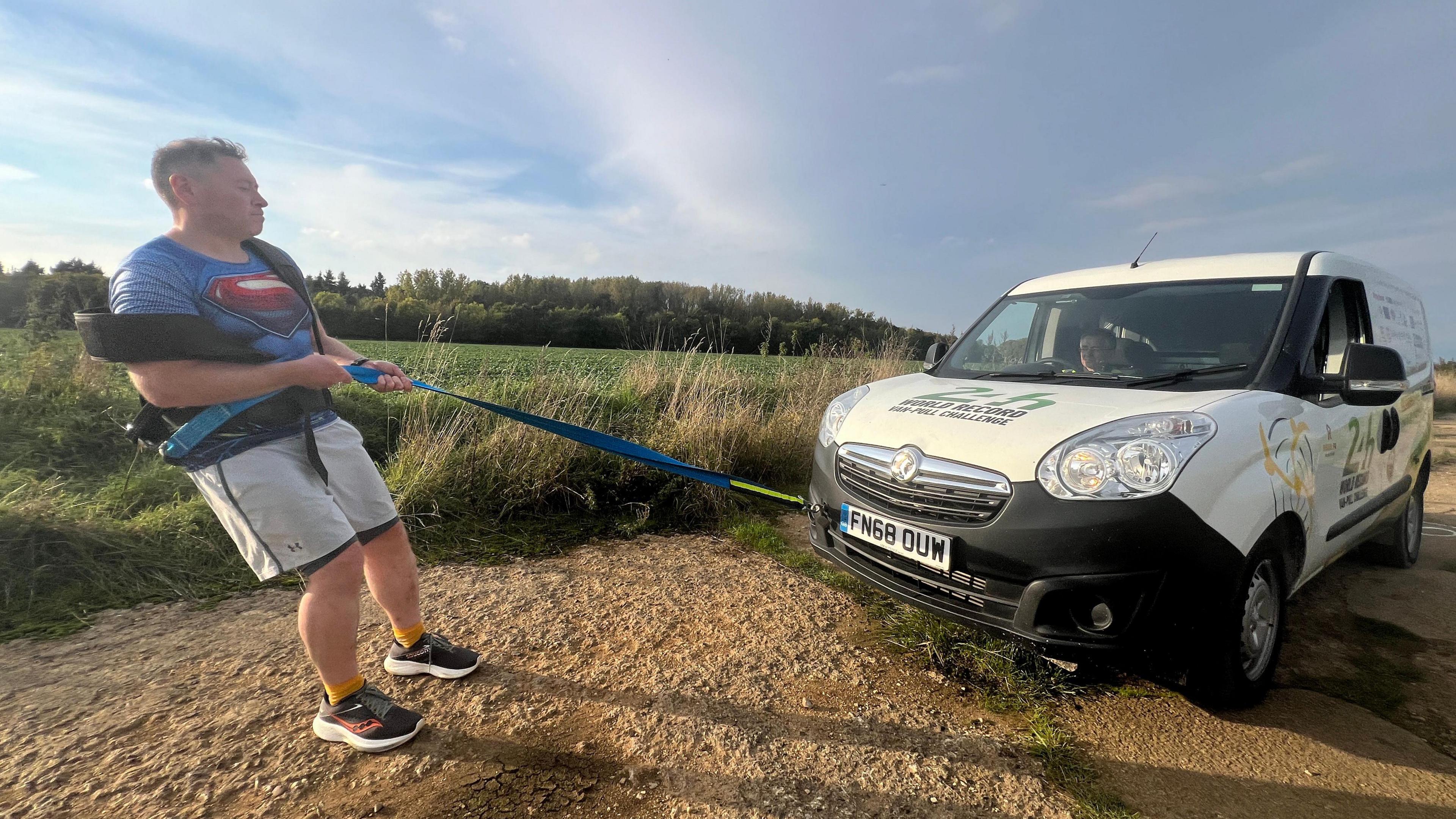 Mike Land pulling a white van on a concreted track, which has lumps and bumps on it, surrounding by green fields and blue sky. He is wearing shorts, trainers, superman t-shirt and a harness for pulling.