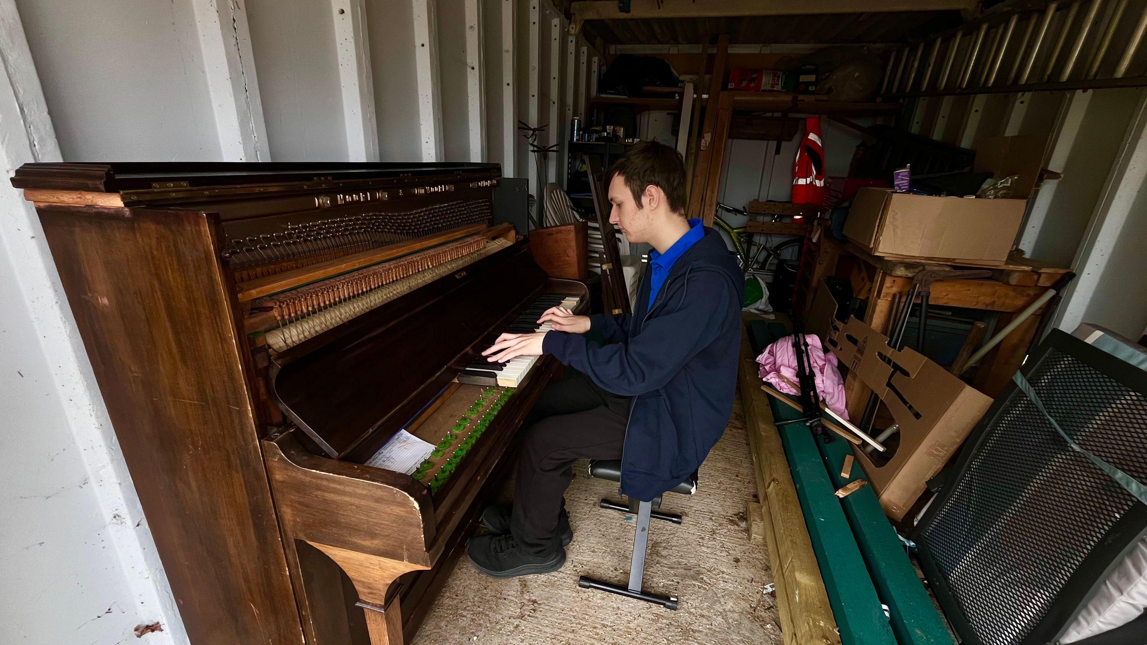 Jamie is sitting in a storage unit, on a stool, playing the broken piano. There is various other bits of junk in the unit. He is wearing a blue polo shirt and a navy blue hooded top.