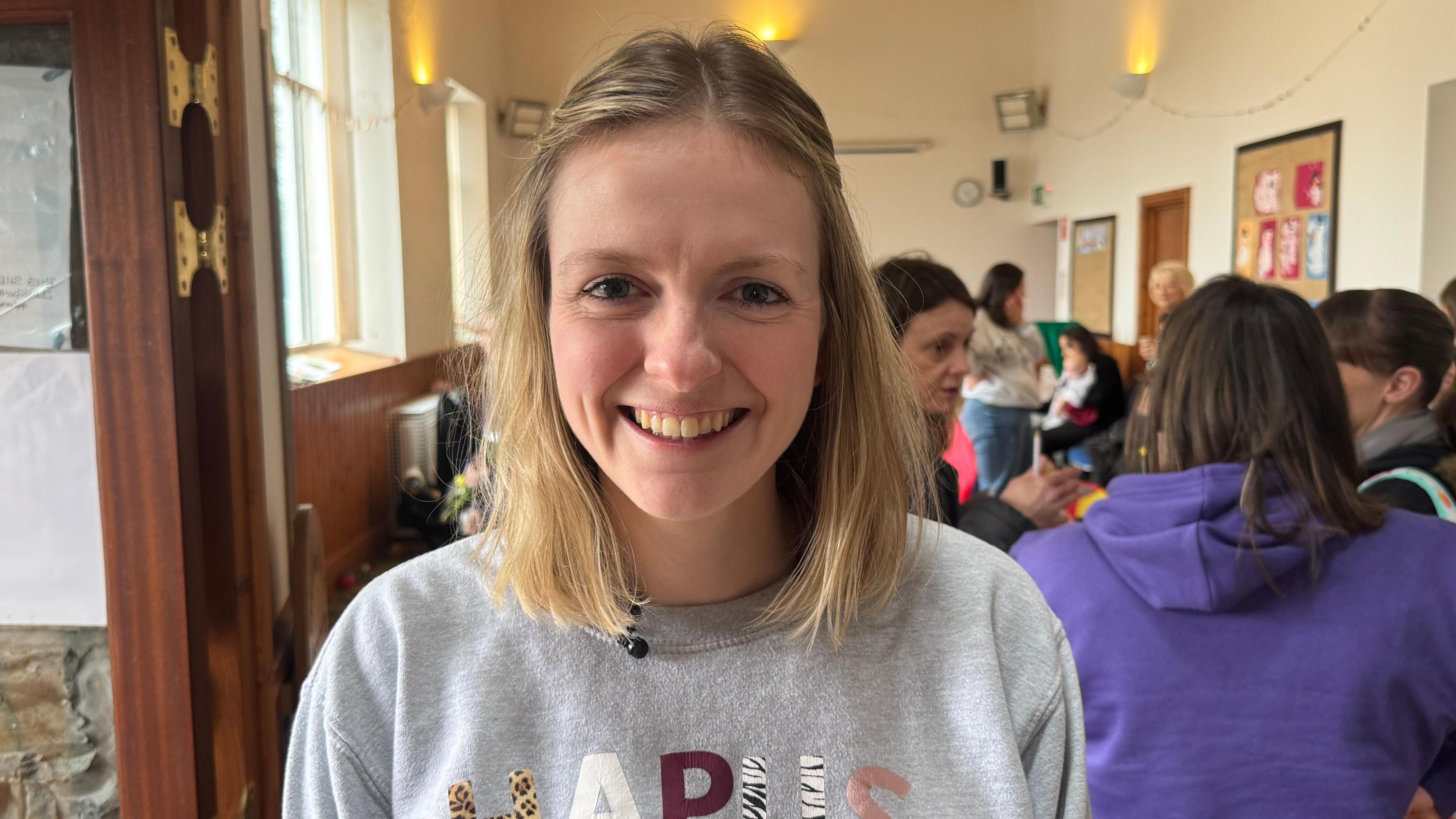 Dynes gyda gwallt melyn yn gwenu o flaen llond neuadd o blant a rhieni. 