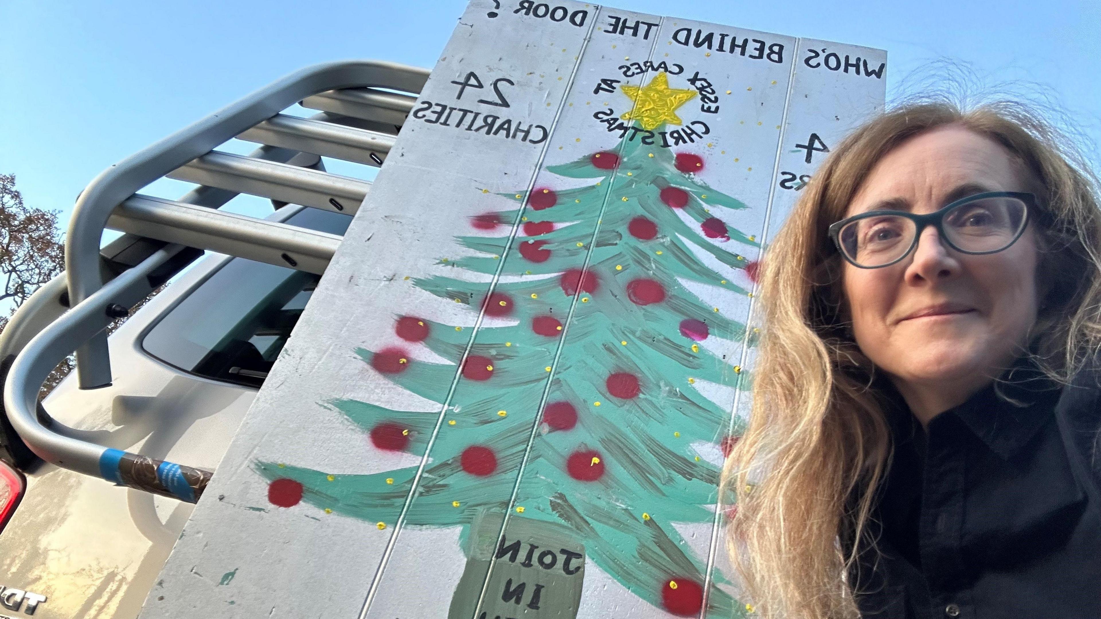 Mary-Ann Deasy has brown hair and is wearing glasses. She is stood next to a large sign with a Christmas tree painted on it. The sign is being used to promote her advent calendar. 