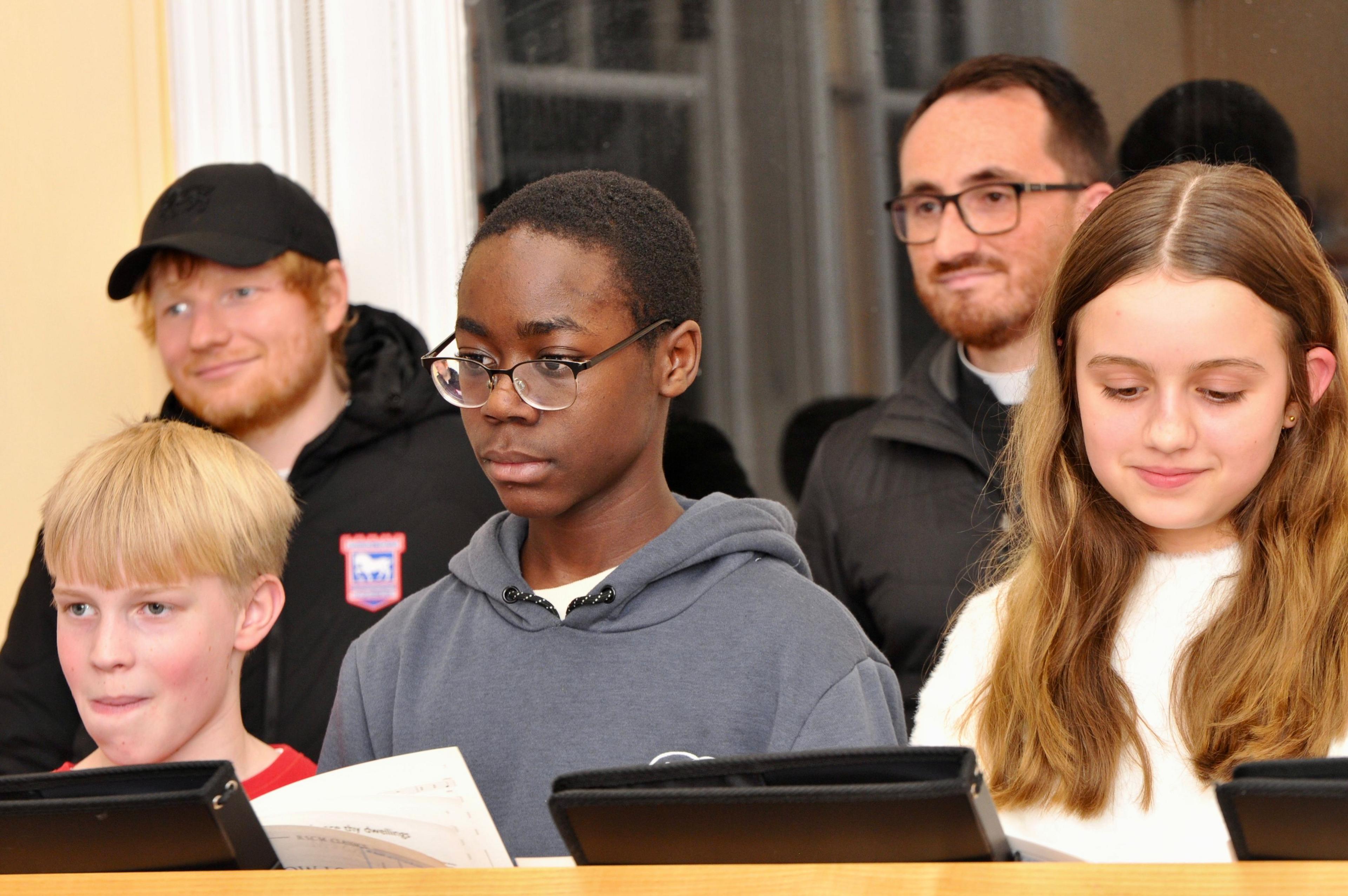 Ed Sheeran indoors wearing a black cap and black Ipswich Town jacket On his left is a vicar, wearing back with a dog collar and with glasses and dark hair and short beard. In front of them are two boys and a girl, who are looking towards music on stands