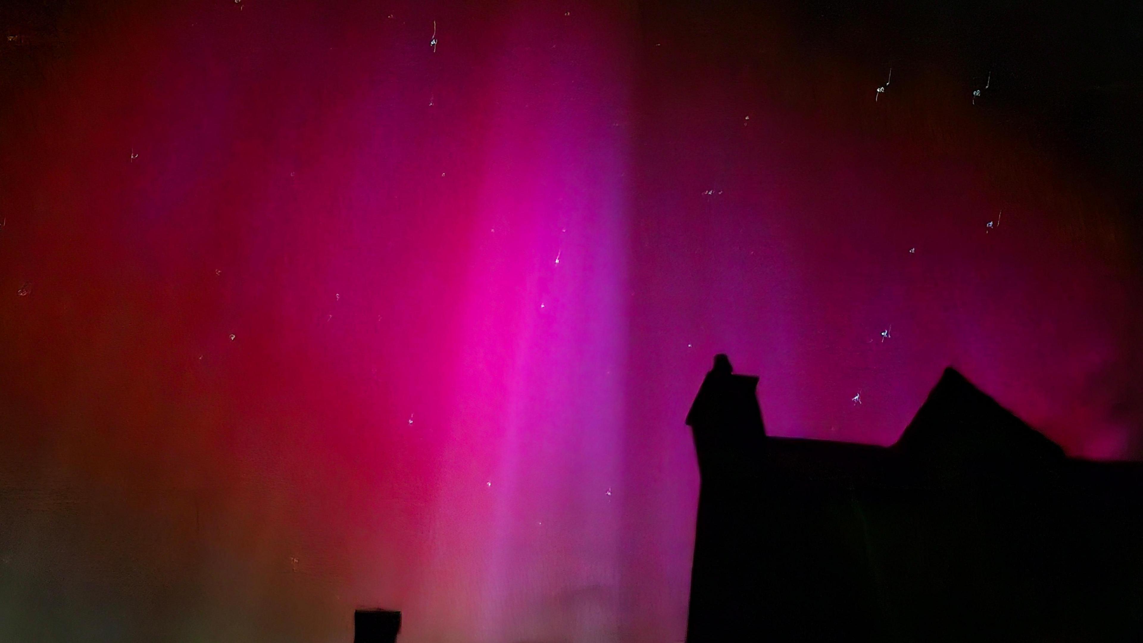 Silhouette of a house roof with very bright colours of the aurora in the sky behind it. Deep purple and red streaks in the sky