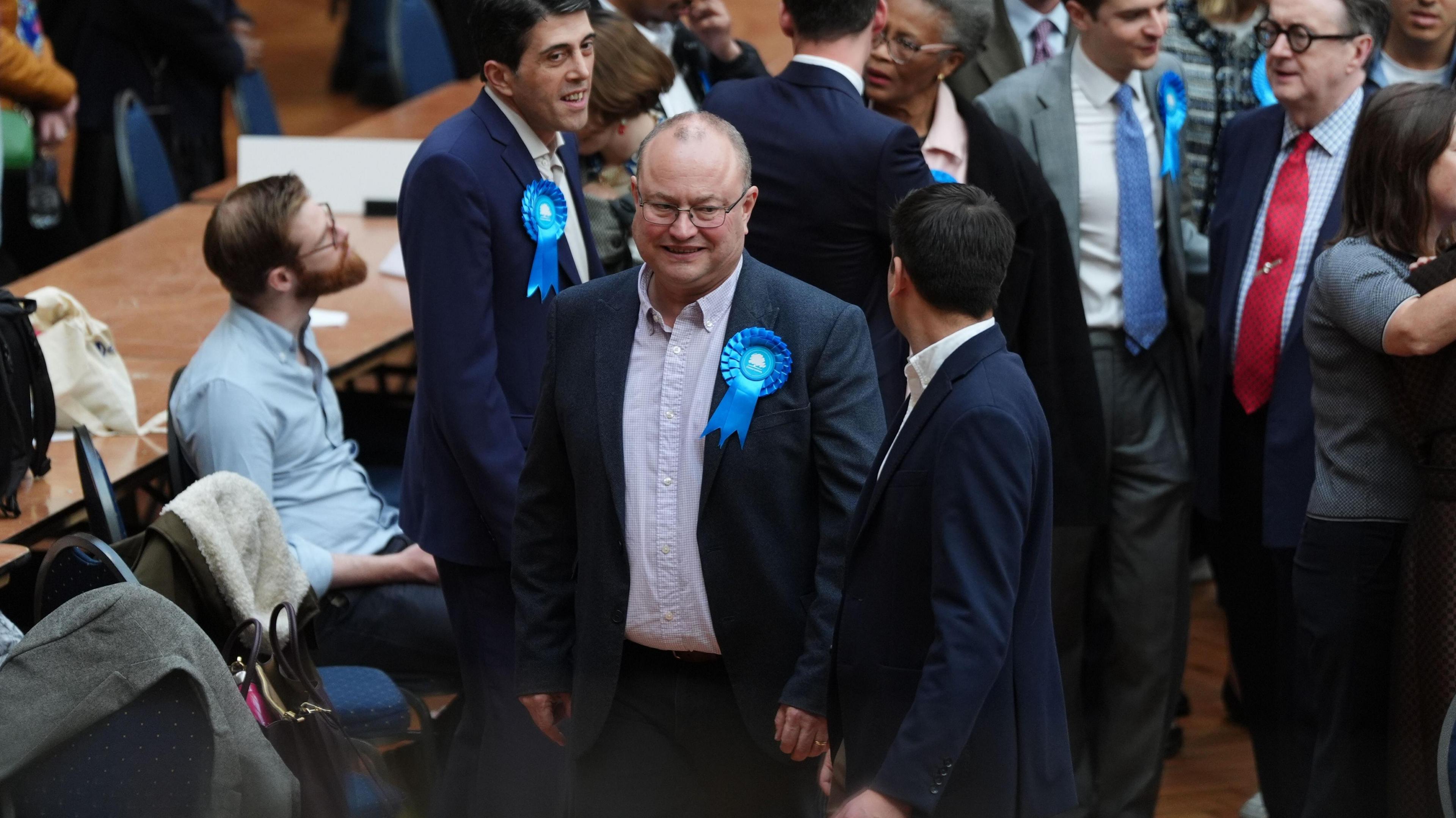 a man wearing glasses and a blue conservative rosette surrounded by other men in suits