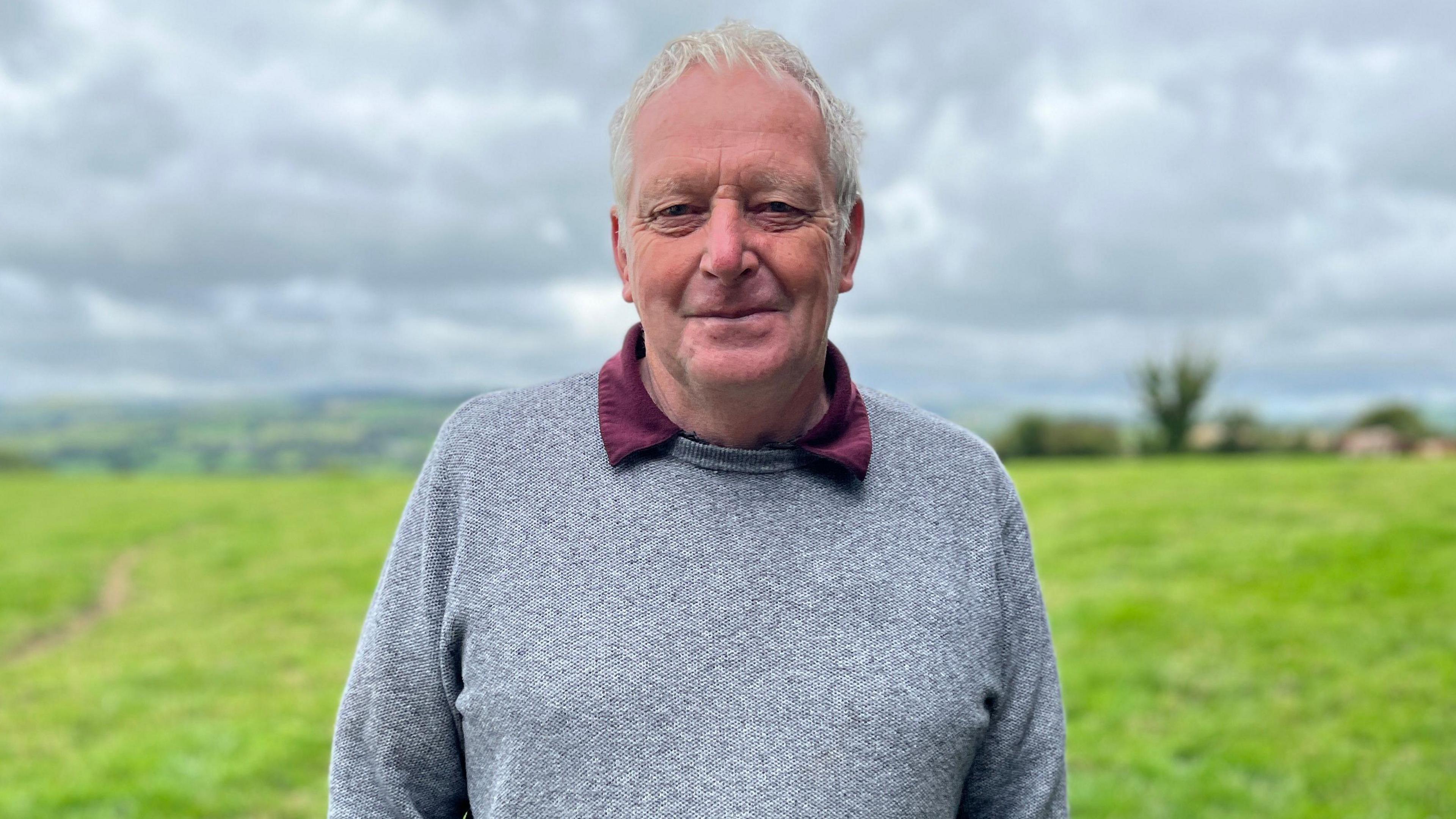 Dyn gyda gwallt gwyn mewn siwmper lwyd yn sefyll mewn cae. 