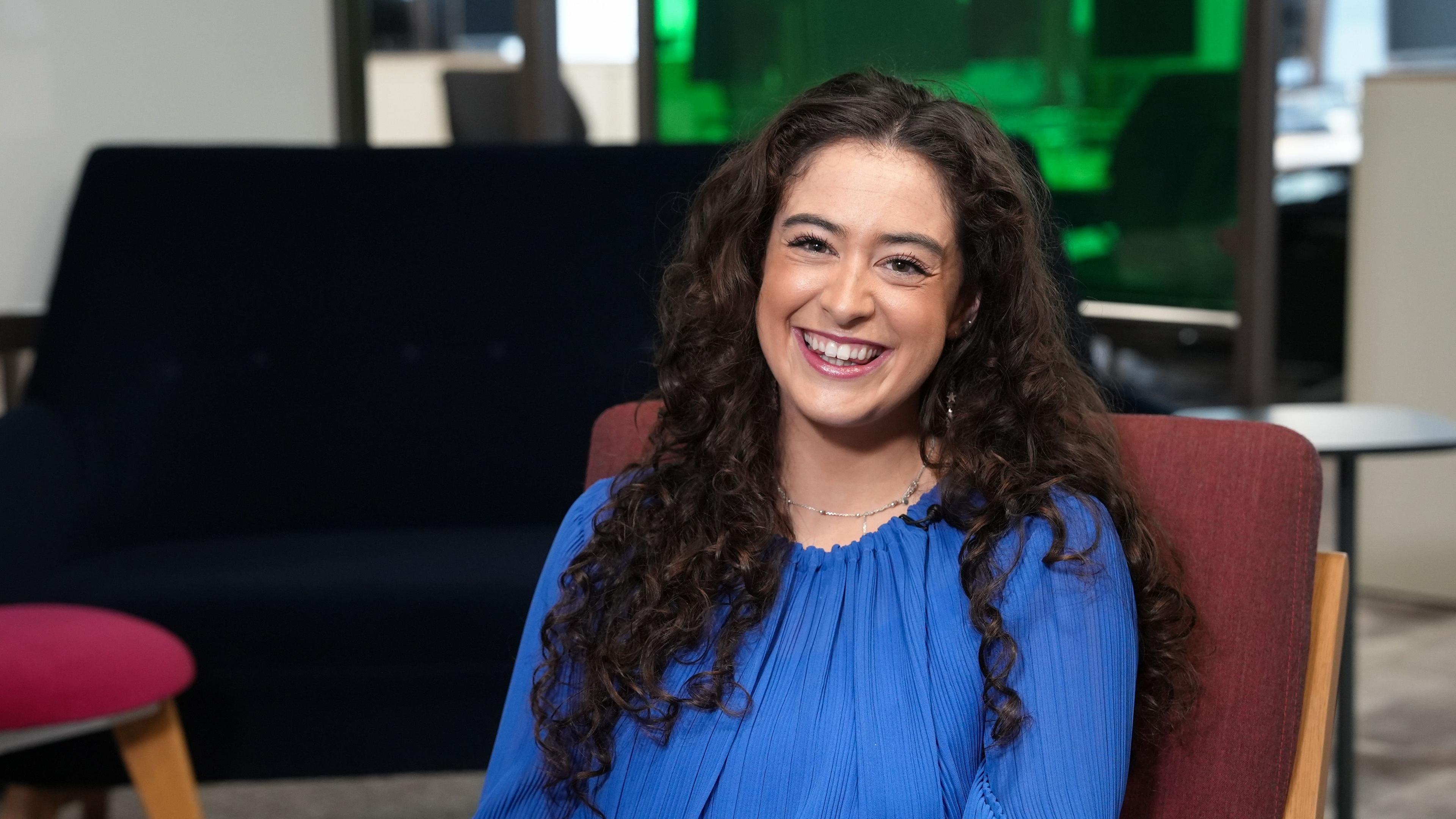 Yn y llun yma mae Elin yn eistedd ar gadair mewn swyddfa. Mae ganddi wallt hir brown a chyrliog a mae hi'n gwisgo crys glas. Mae hi'n gwenu.