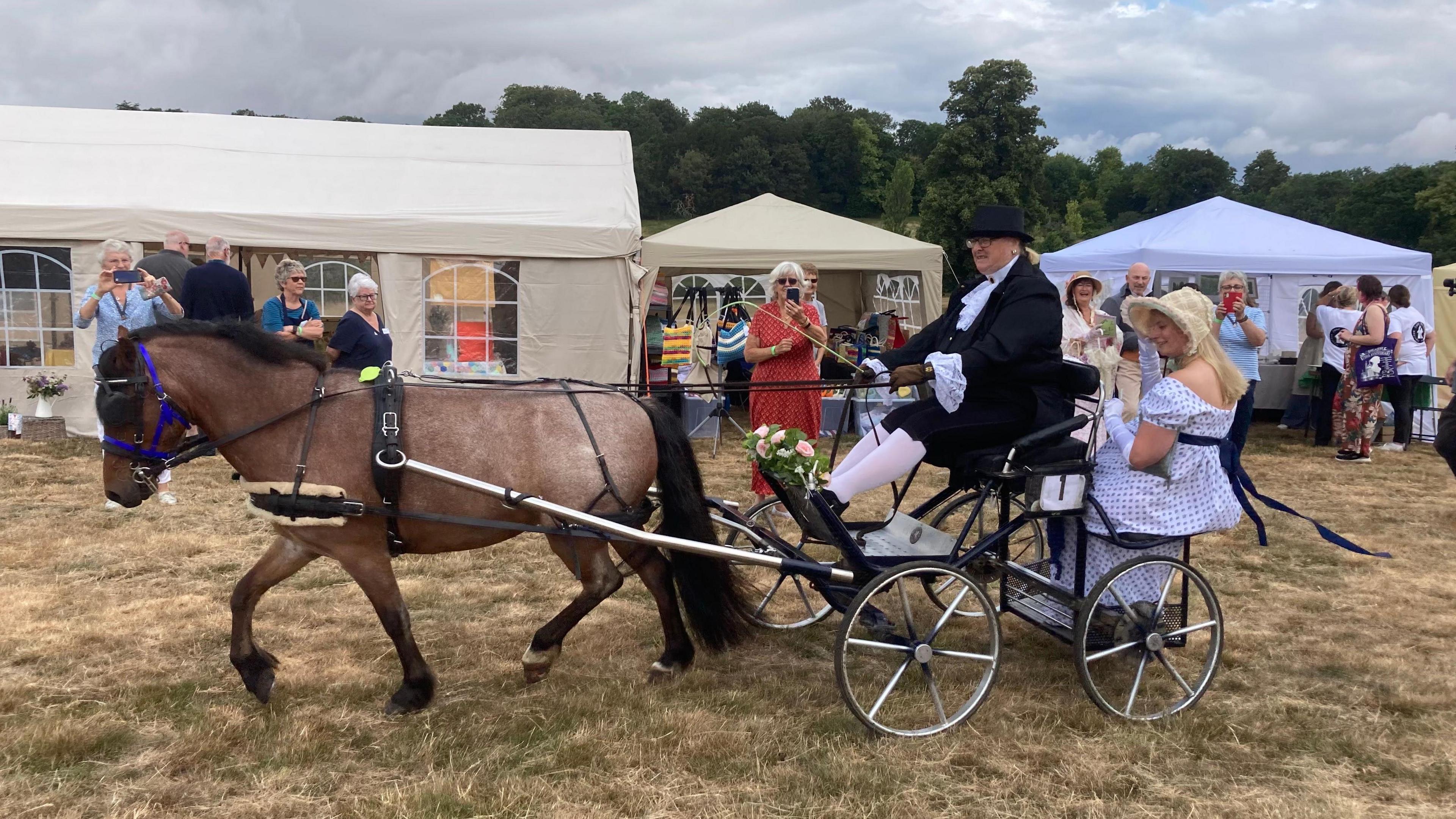 Horse pulling cart with man driving and woman in regency dress sat in the buggy.