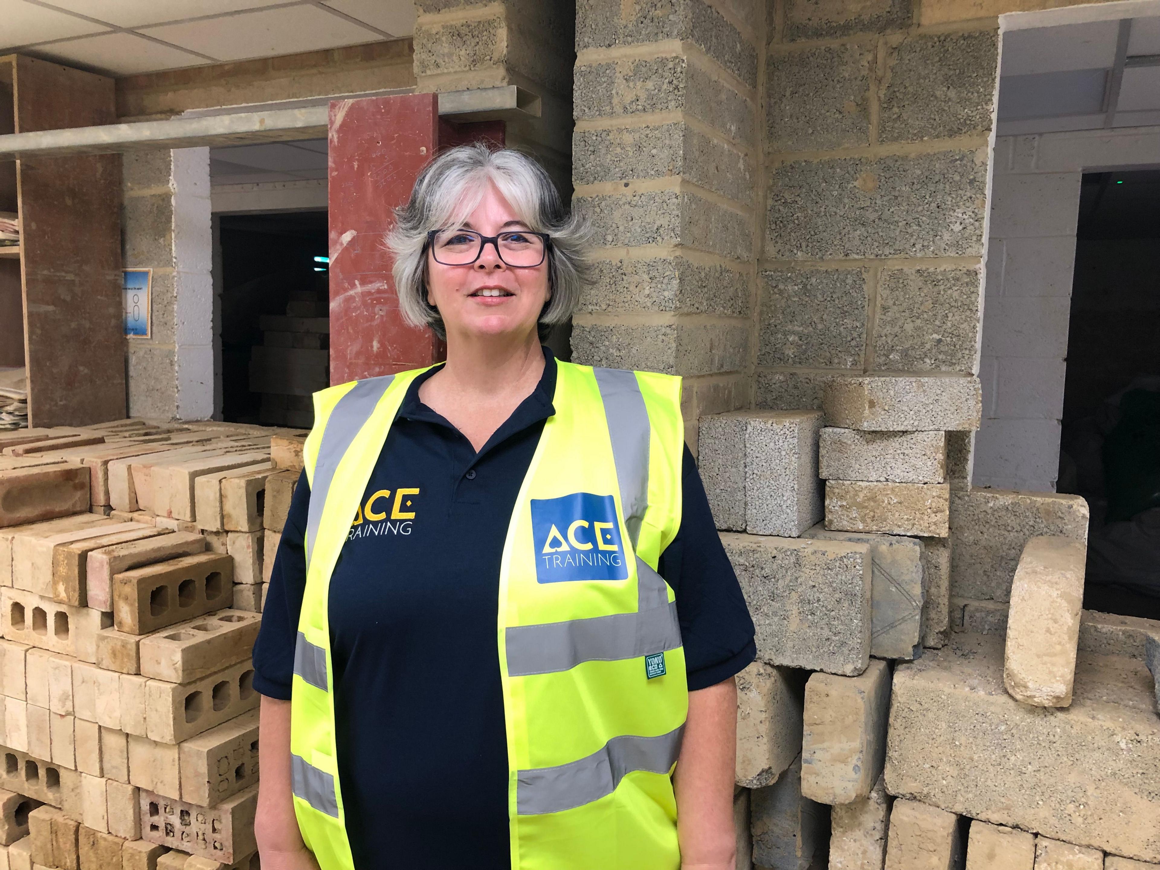 Natalie Games stands in front of a large pile of bricks and breeze blocks. She is wearing a short-sleeved black top and a sleeveless high-vis jacket bearing the ACE logo in blue. She has grey hair cut in a long bob with a fringe and is wearing glasses.