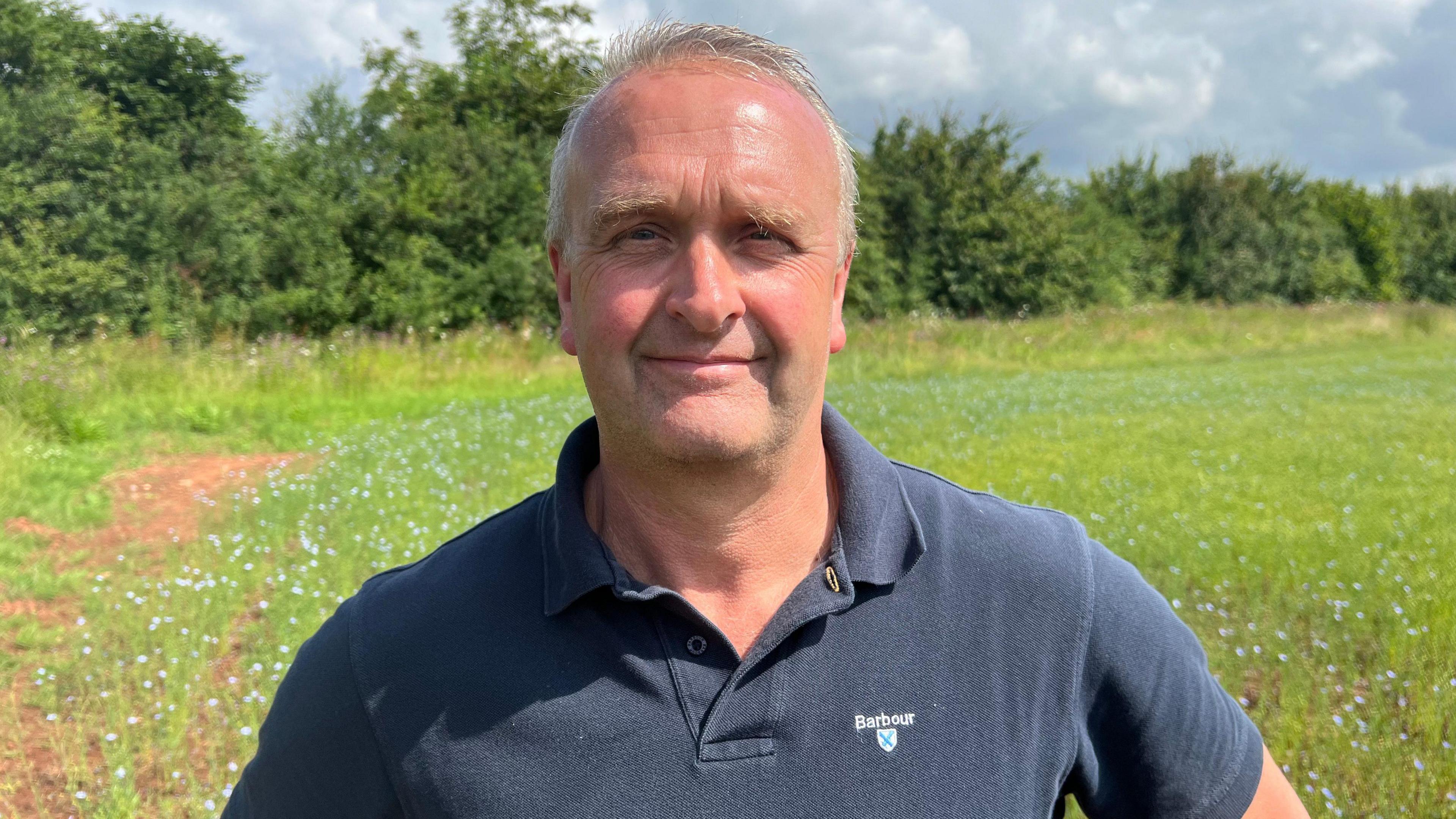 Jeremy wearing a blue polo t-shirt standing in front of his farm