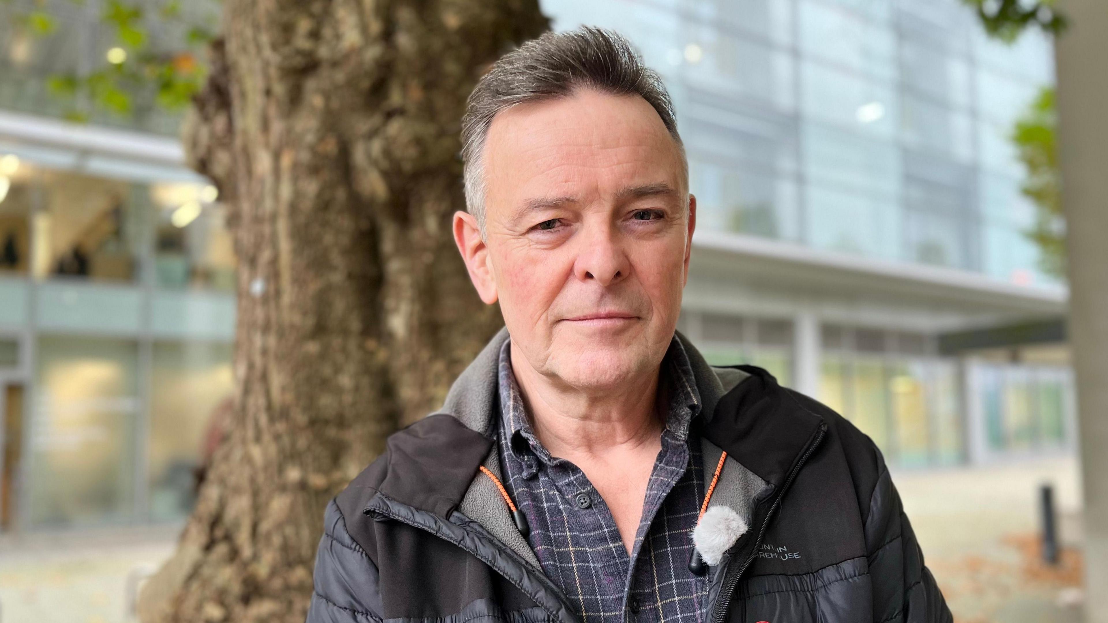 Ken McBride has short greying hair. He is wearing a checked blue shirt with a hooded jacket. There is a tree trunk and an office building behind him.