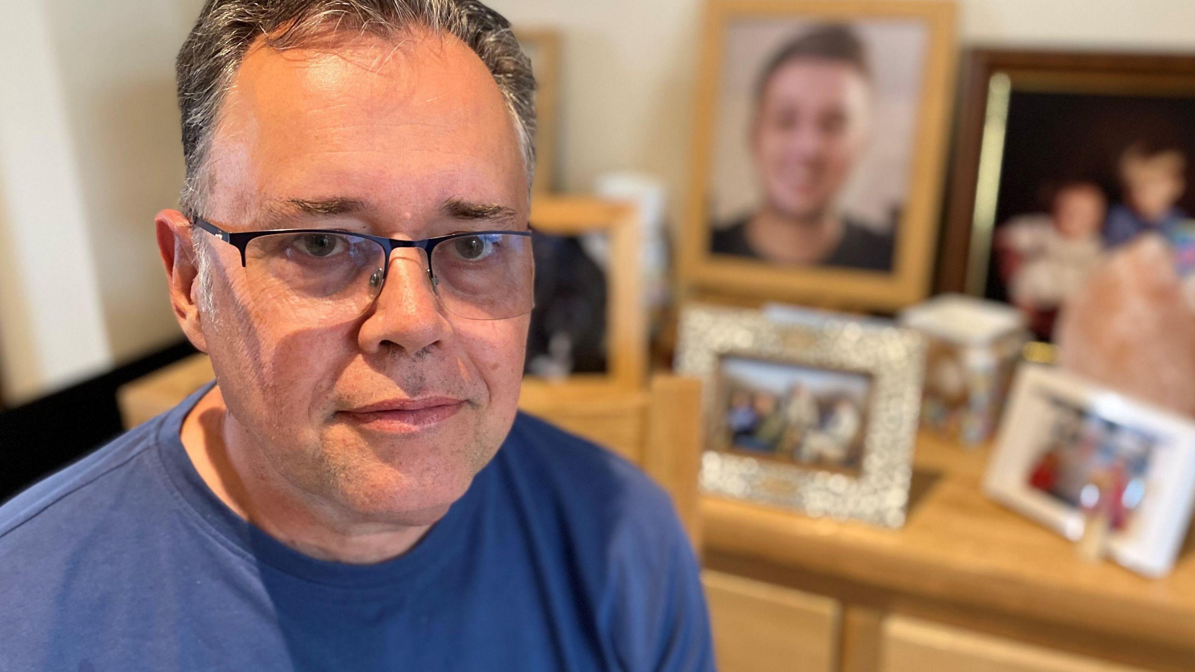 Mark Pegram sitting in a room with family photographs including ones of his son Sam behind him. He is wearing glasses and a blue top. 