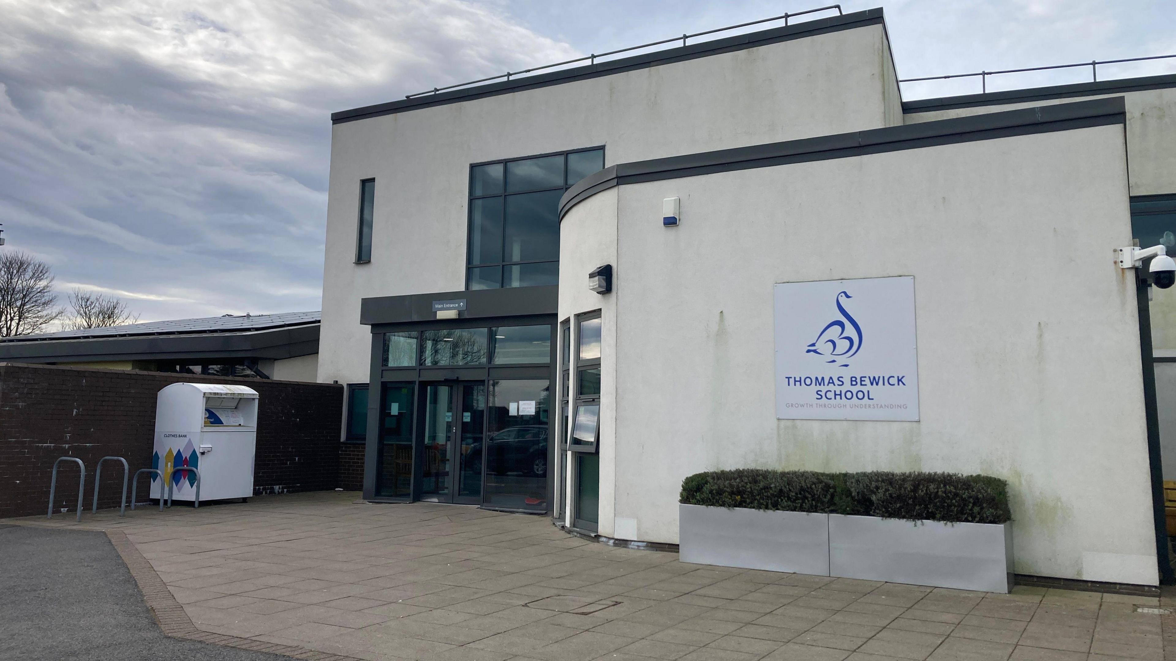 The entrance to Thomas Bewick School which is a small building with white concrete walls and windows painted brown. To one side are bike racks and a recyling container