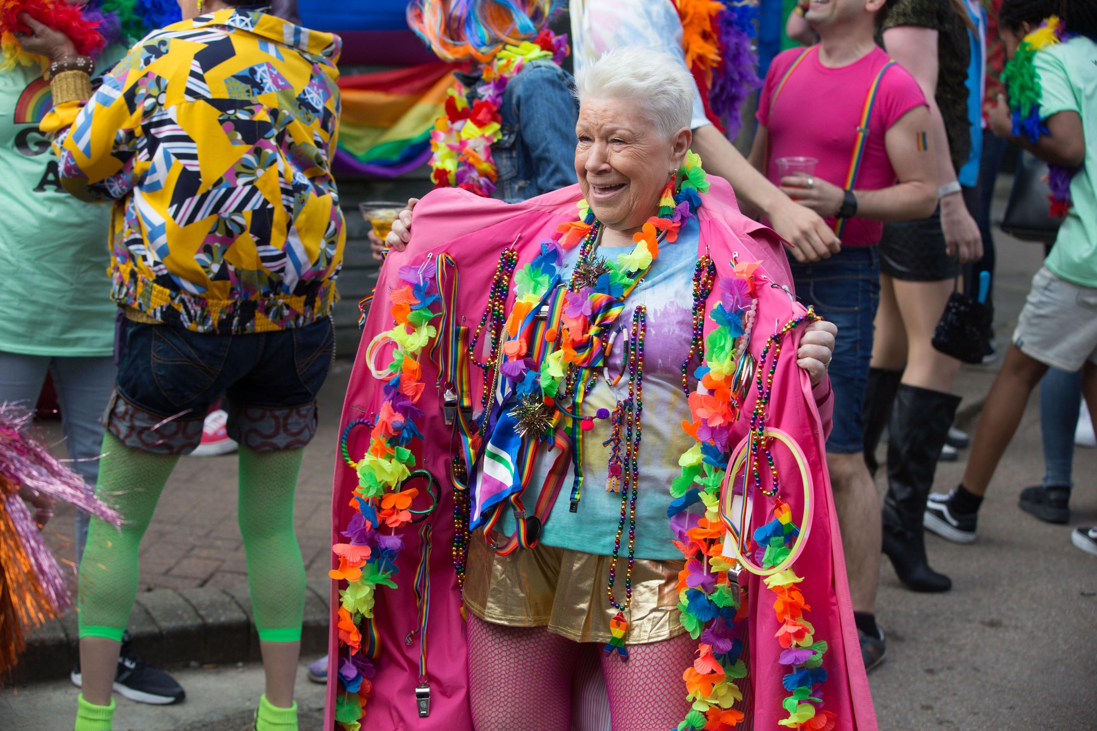 picture of little mo slater, an old woman with short white hair in a tye-dye tshirt, gold hotpants and red fishnets tights. she is opening up her pink coat, revealing multiple rainbow coloured items attached to the inside with safety pins