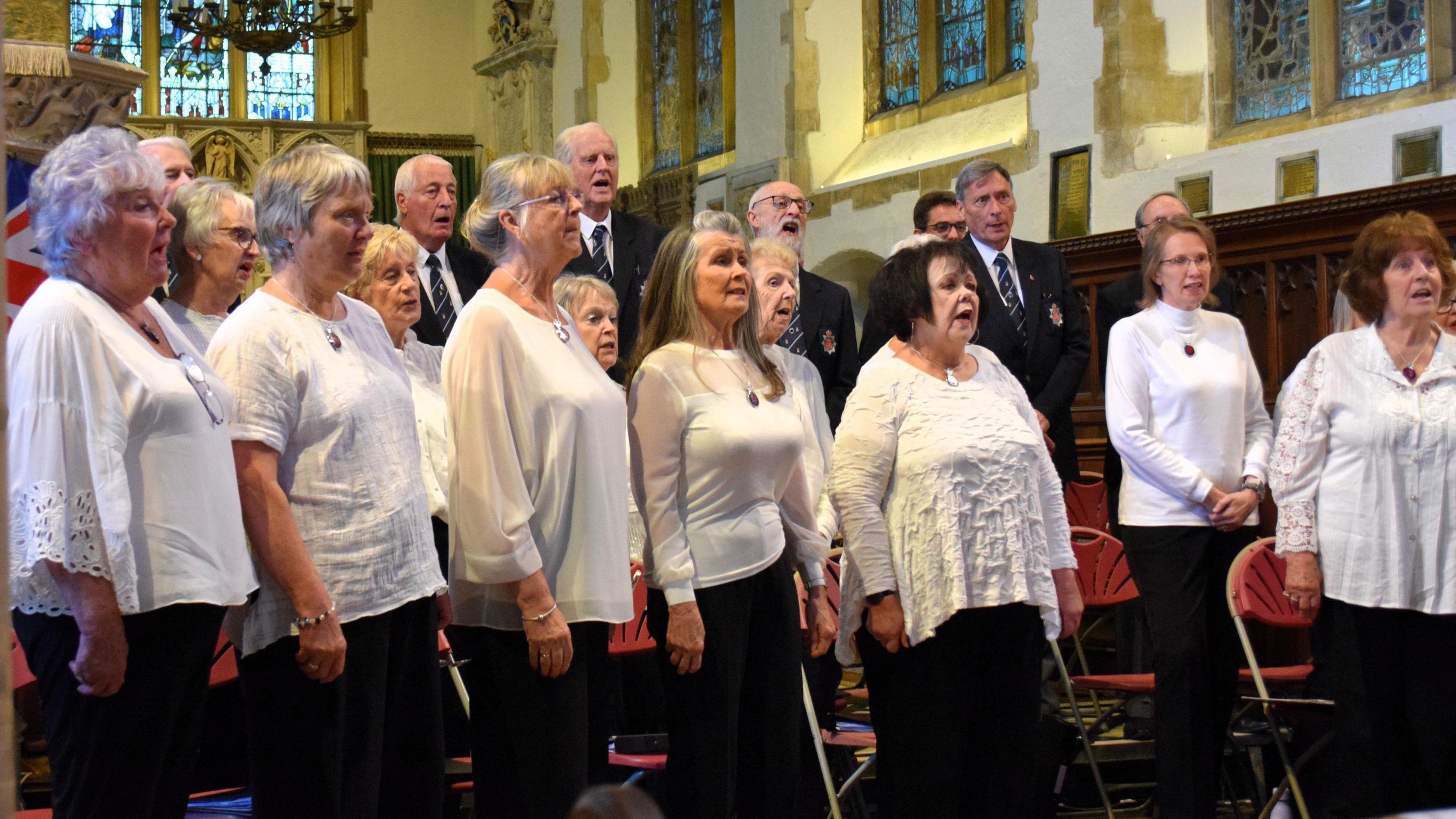 Members of the Essex Police Community Choir singing in a church.  they are standing up.  The interior walls are cream and white and there are stained glass windows.