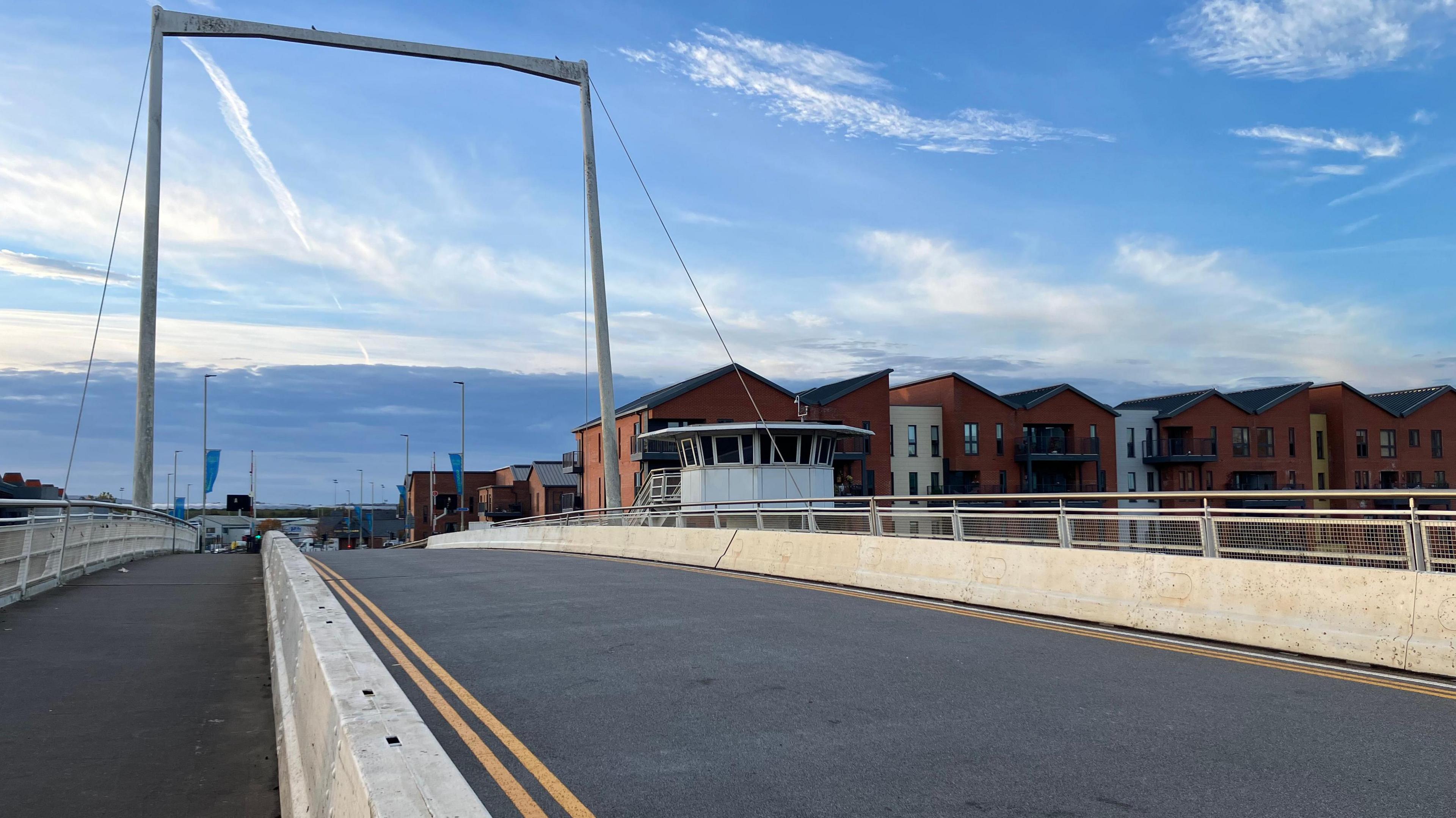 The new resin surface, with double yellow lines, on the road of High Orchard Bridge in Gloucester. There is a white supportive arch over the top of the bridge, which goes over the canal. New build flats and a watchtower can be seen behind the bridge on an autumn evening as the sun sets.