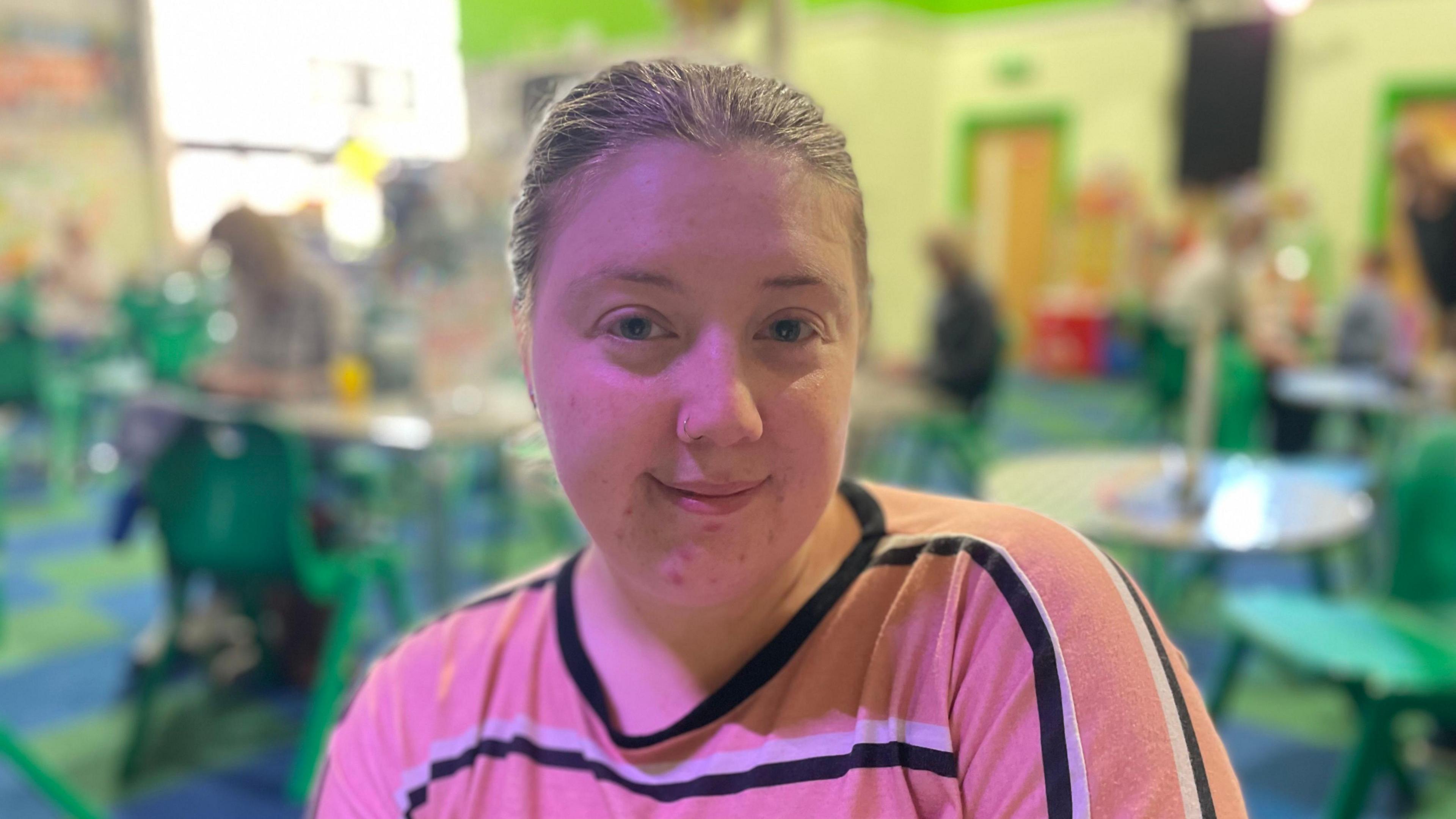 A woman smiling and looking directly into the camera. She is wearing a pink, black and white striped t-shirt. There are tables and chairs in the background.