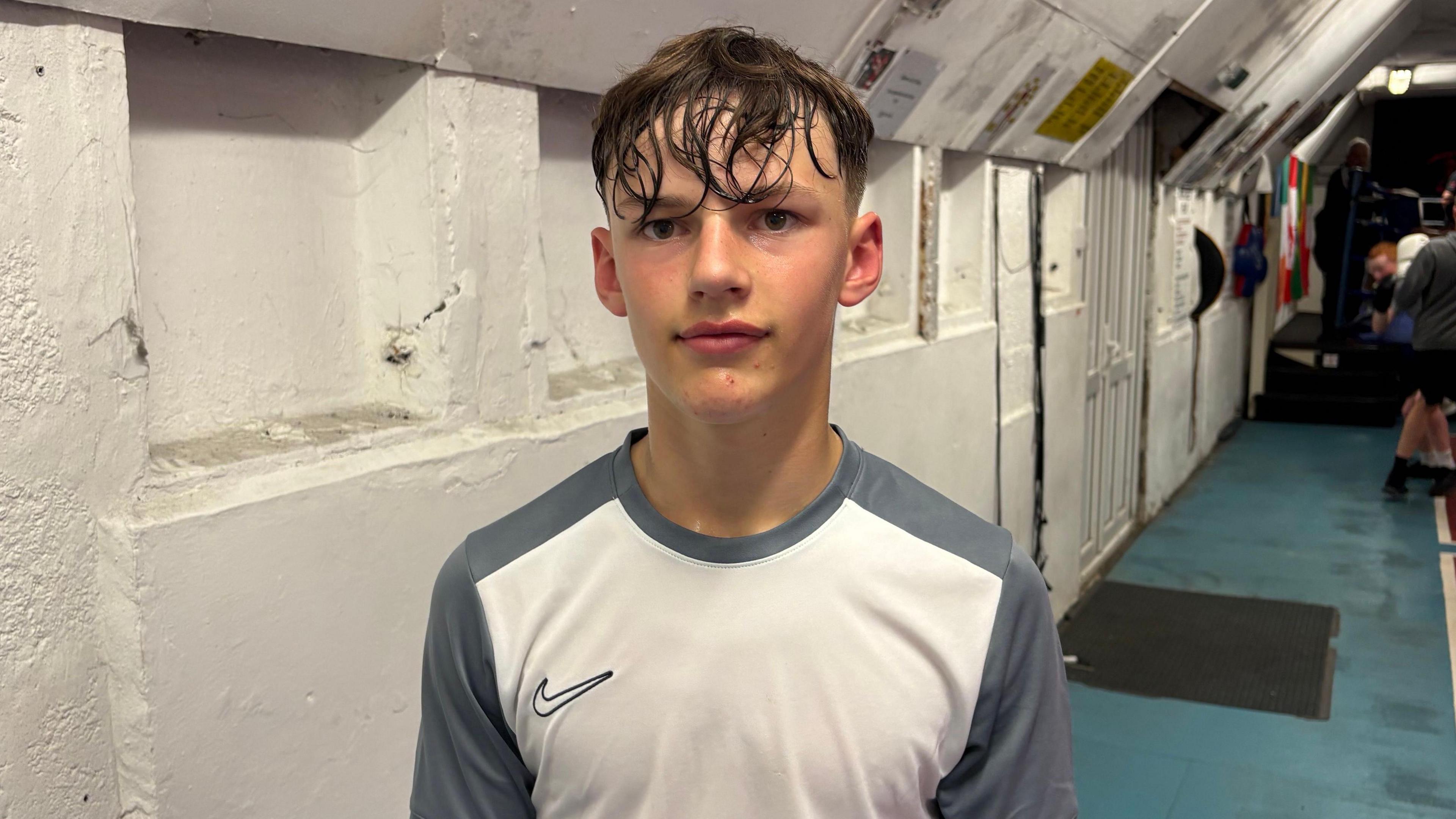 A young lad with sweaty dark hair wearing a grey and white Nike training top standing in front of a white wall 