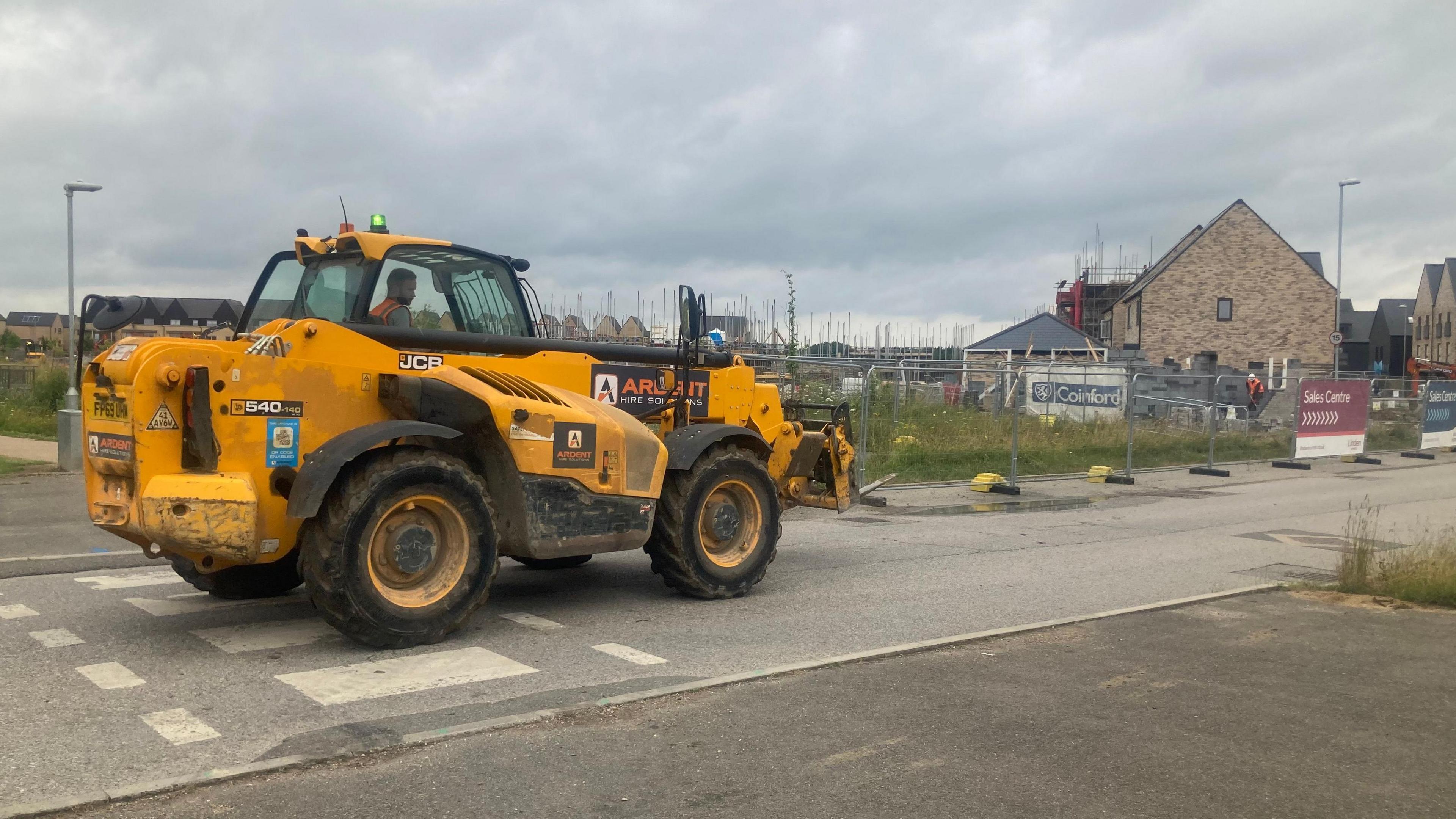 A yellow JCB digger at the entrance to a housing construction site, with a row of partially-built houses in the background