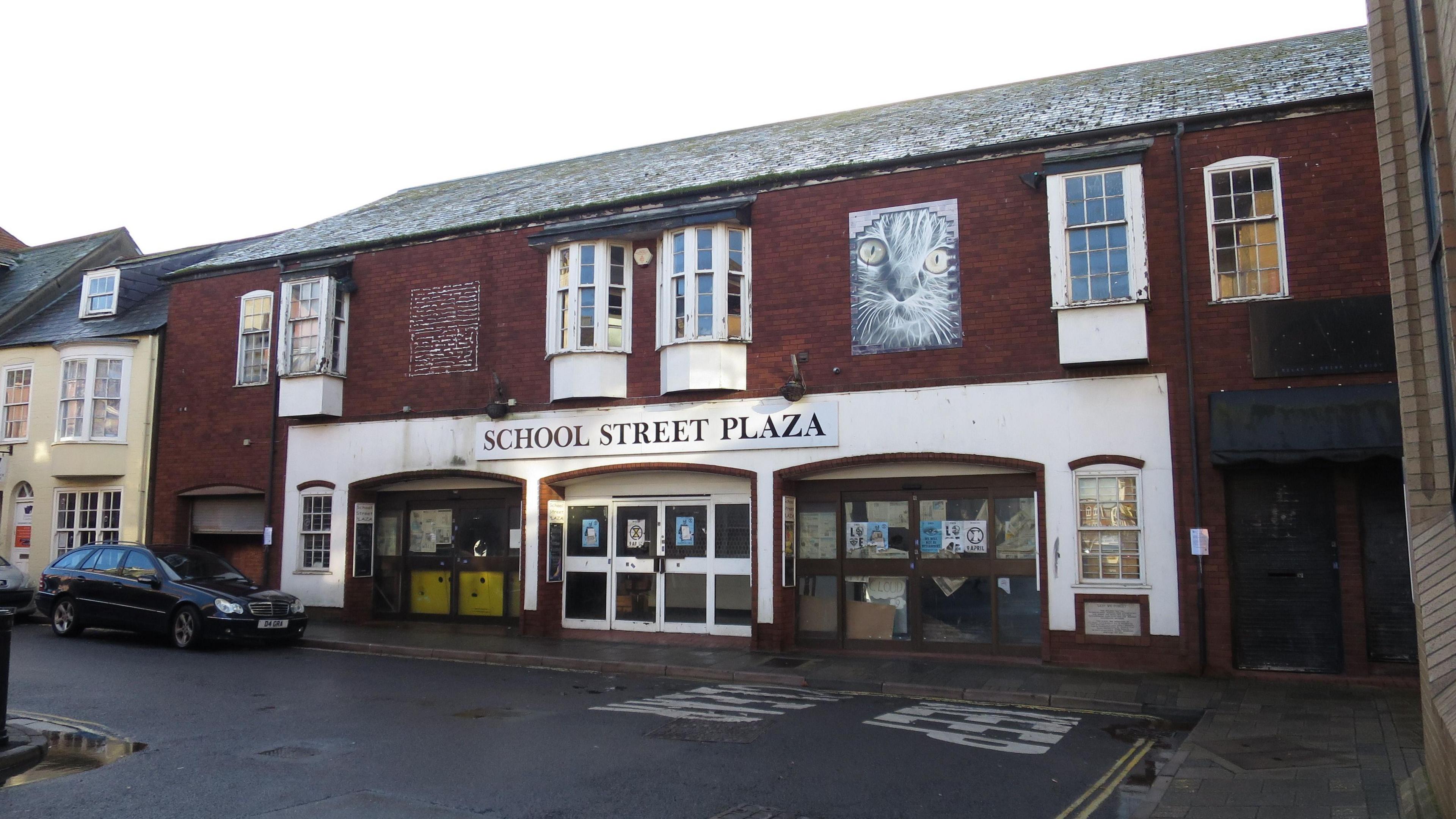 Exterior or School Street Plaza - a two-storey brick building in a row of terraced houses. The frontage takes up the space of about three or four terraced houses. There are double doors in the centre and two units either side with their own entrances on to the street. The whole building is abandoned with posters and newspaper stuck over the windows.