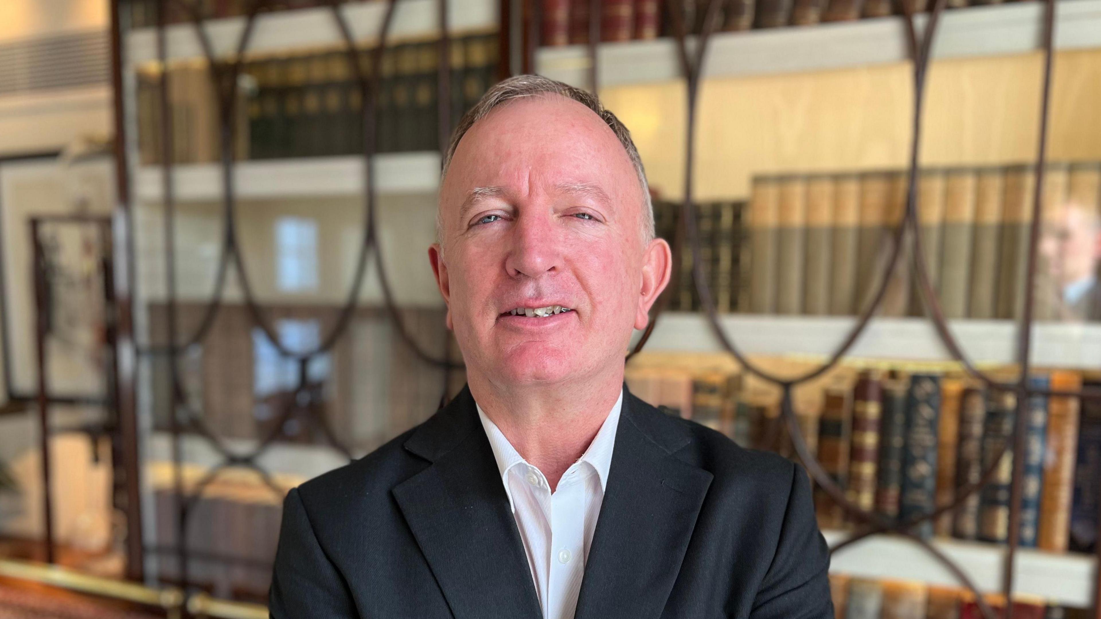 Alan Sillett - A man with wispish grey hair, wearing a black suit over a white shirt.