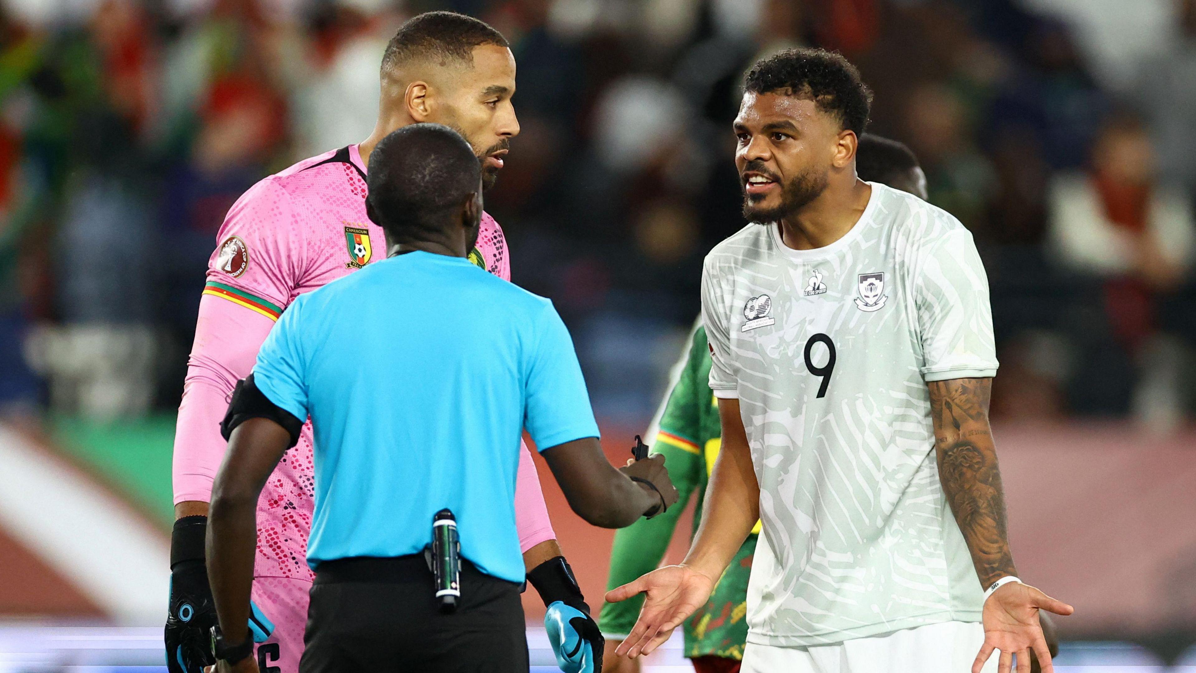 Lyle Foster, wearing a white South Africa shirt with a number nine on the chest, remonstrates with a referee seen from behind and wearing a blue shirt as Cameroon goalkeeper Devis Epassy, wearing pink, is seen standing alongside Foster
