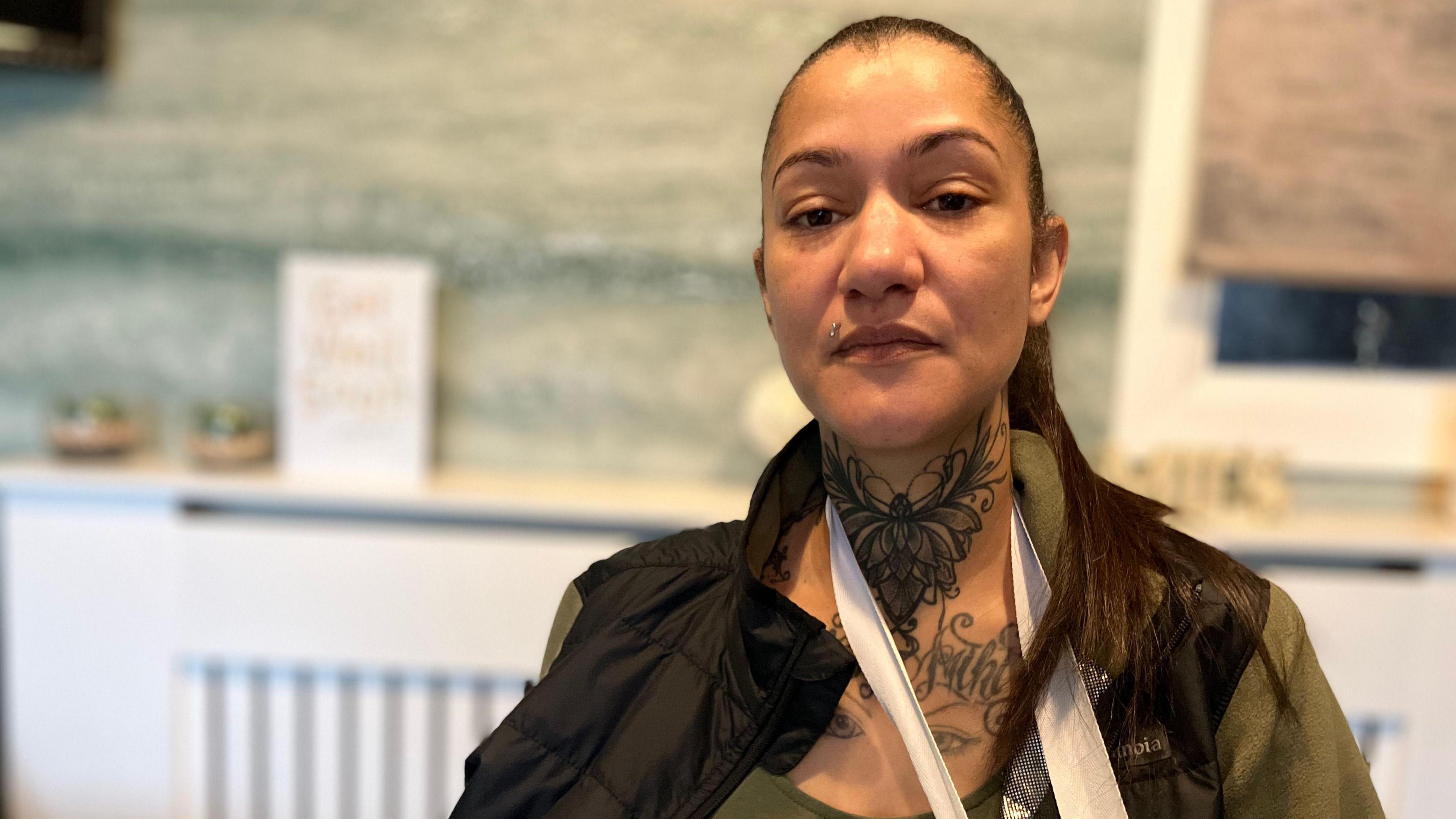 A woman, Michelle Farara, wearing a black body warmer jacket and green shirt. She has a large tattoo of a butterfly on her neck and has a white bandage sling around her neck. She has brown hair pulled back into a long ponytail.