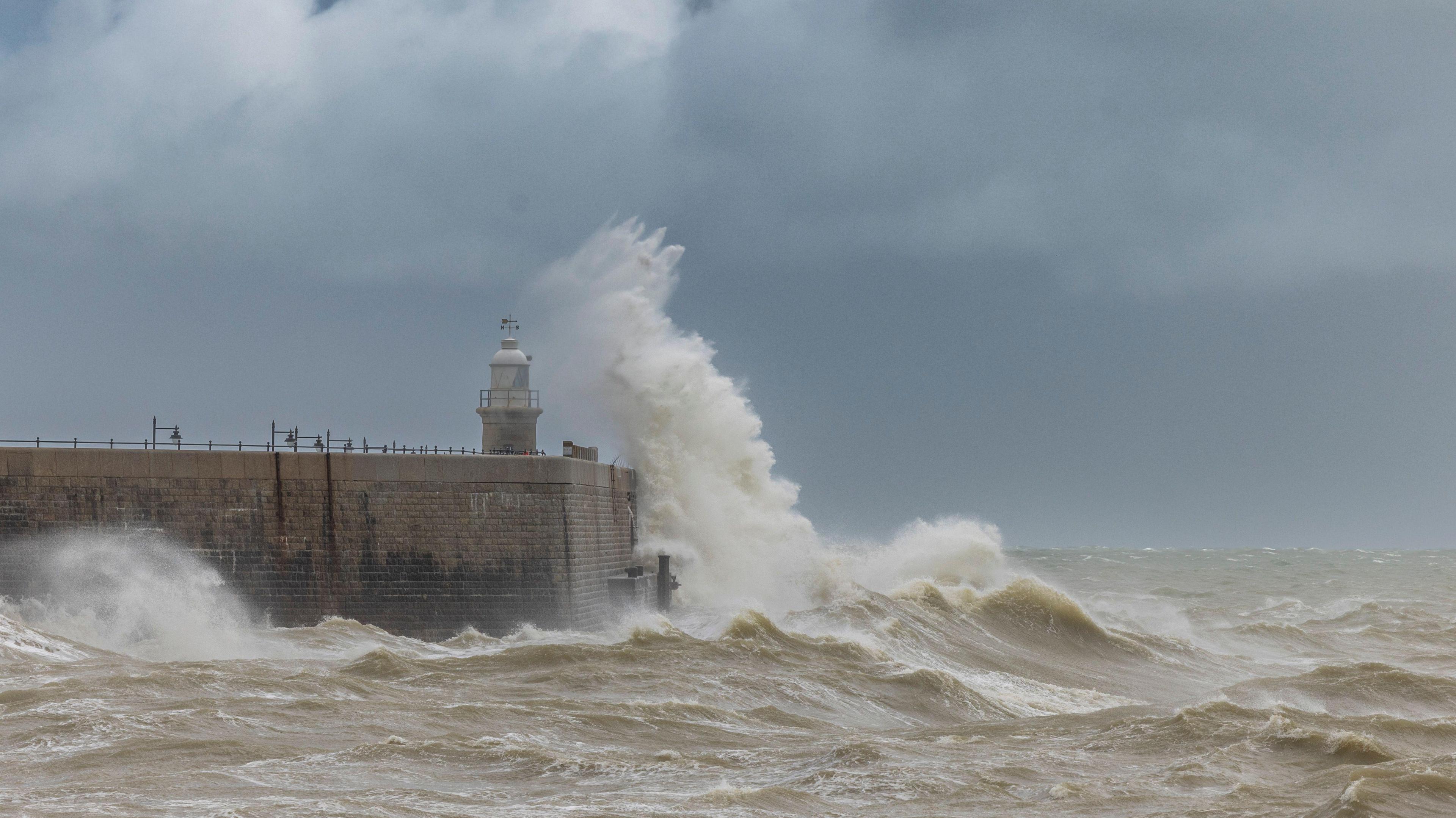 A large wave crashing over a sea wall with a small lighthouse at the end of it, with dark skies and a rough seas