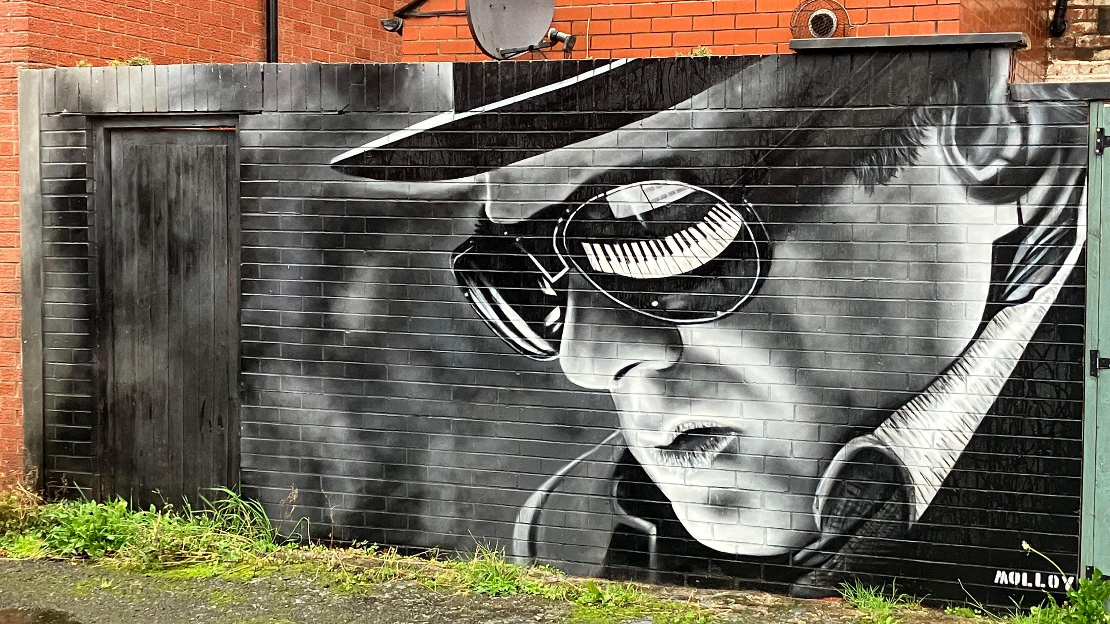 A painted mural of Sir Van on a bricked wall. The wall is painted black. A piano is reflected in Sir Van's sunglasses.
