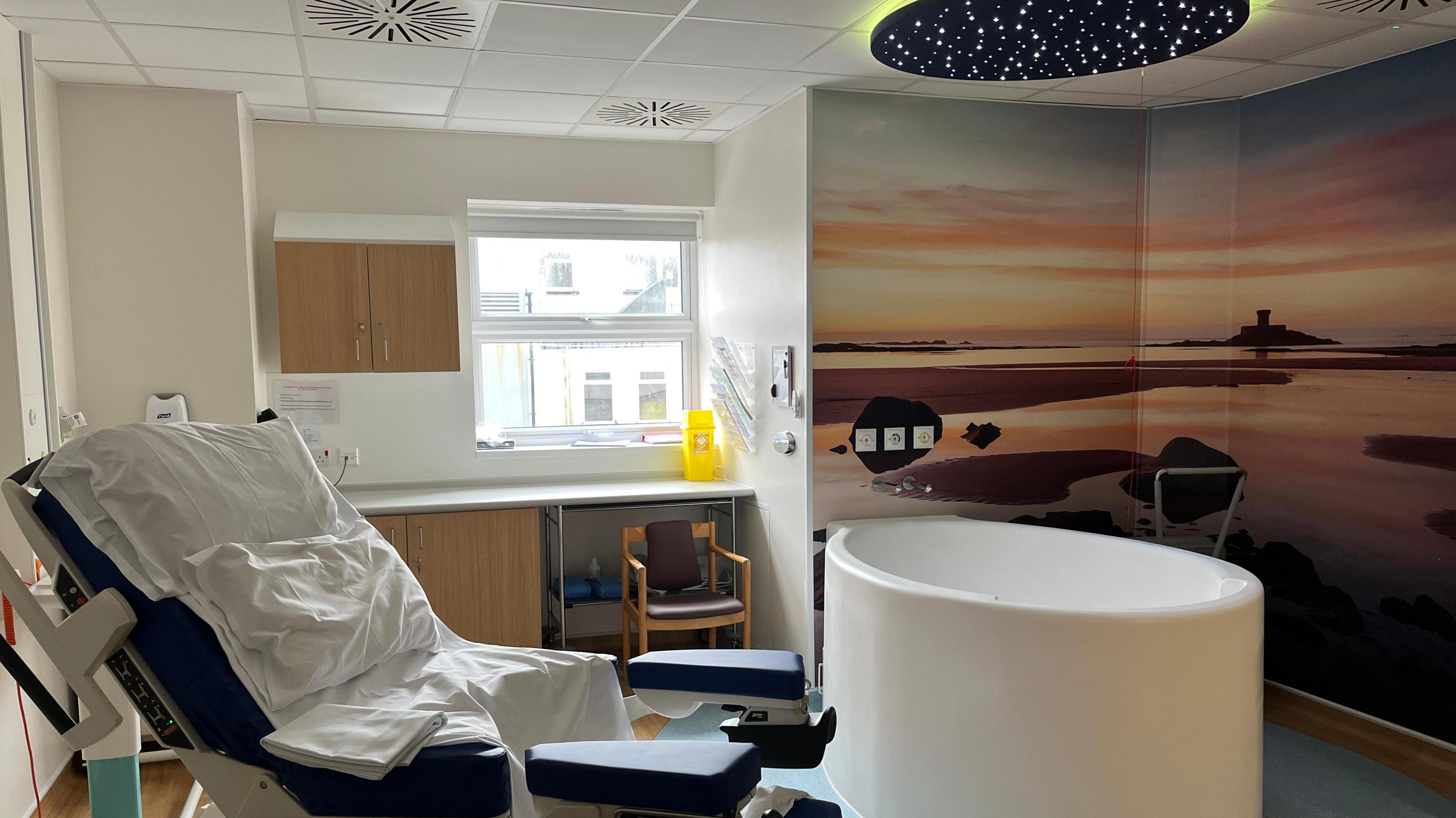 Inside a birthing room at the General Hospital in St Helier, Jersey. There is a hospital chair, a round bath, a sunset image on the wall and a window at the back. 
