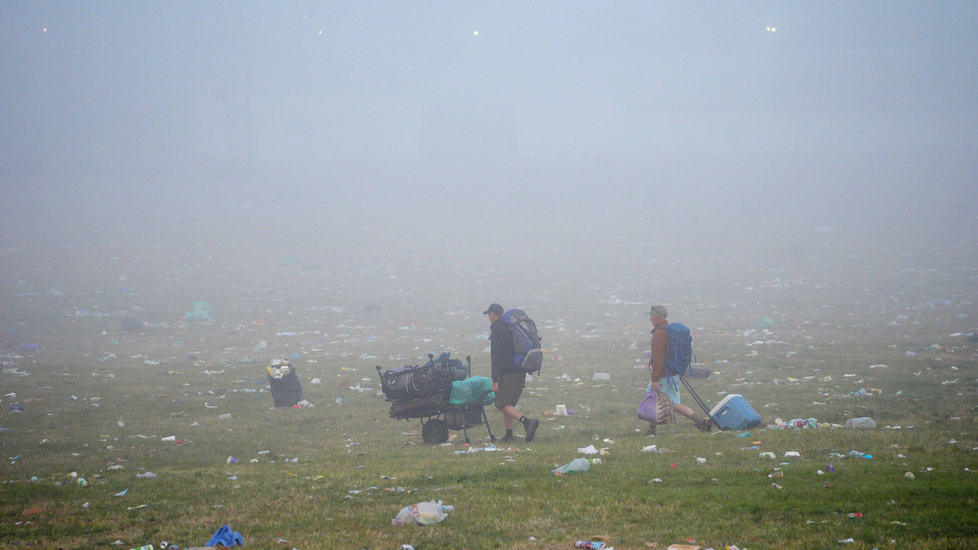 Two punters walking across a misty field at Glastonbury. They are carrying, pushing and pulling their belongings. Lots of litter can be seen all over the field.