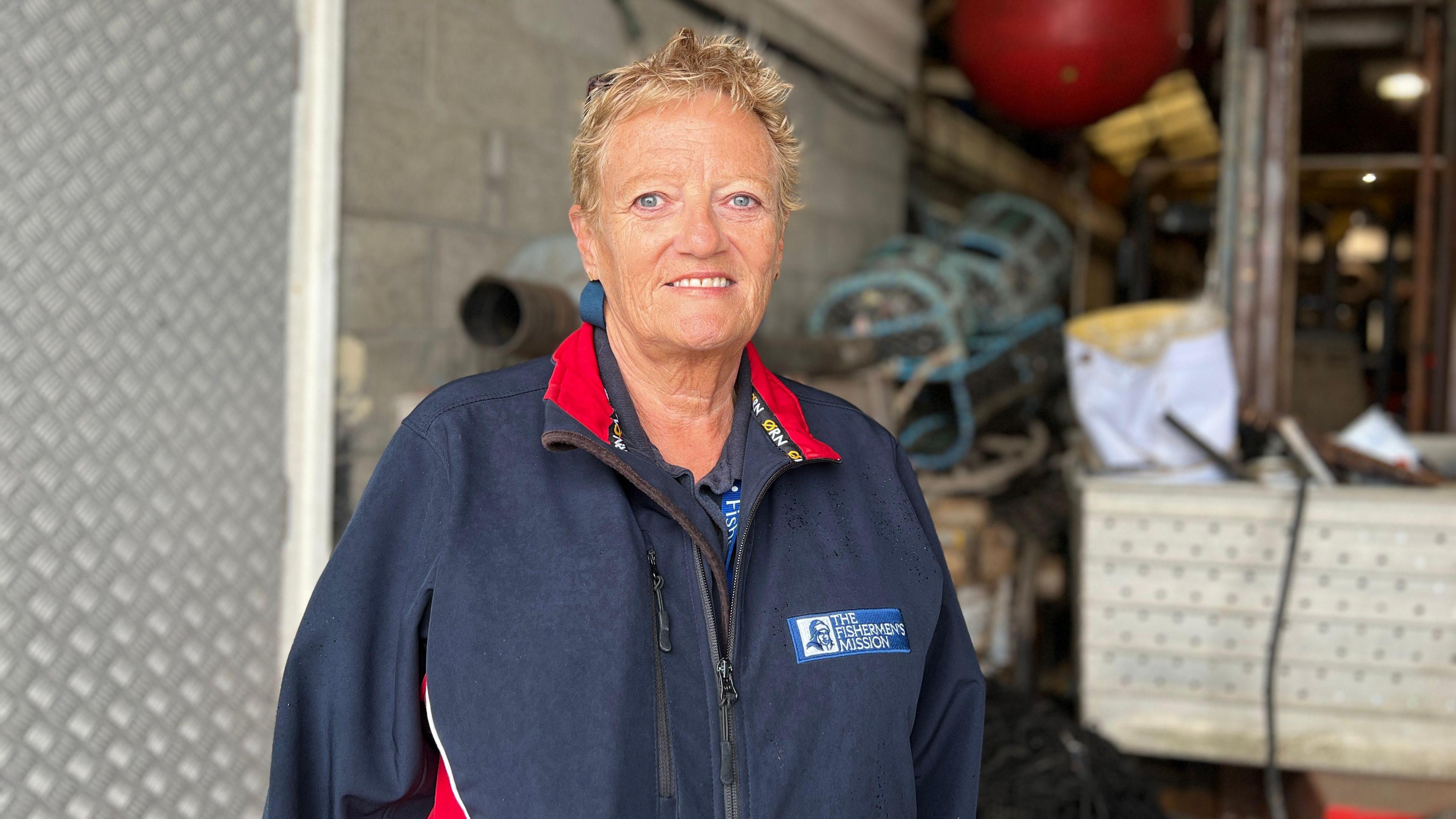 Carol smiles at the camera. She is wearing a blue fleece with red linings and a Fishermen's Mission's logo on the chest. She has short blonde hair and blue eyes. Behind her is a warehouse containing fishing equipment such as nets.