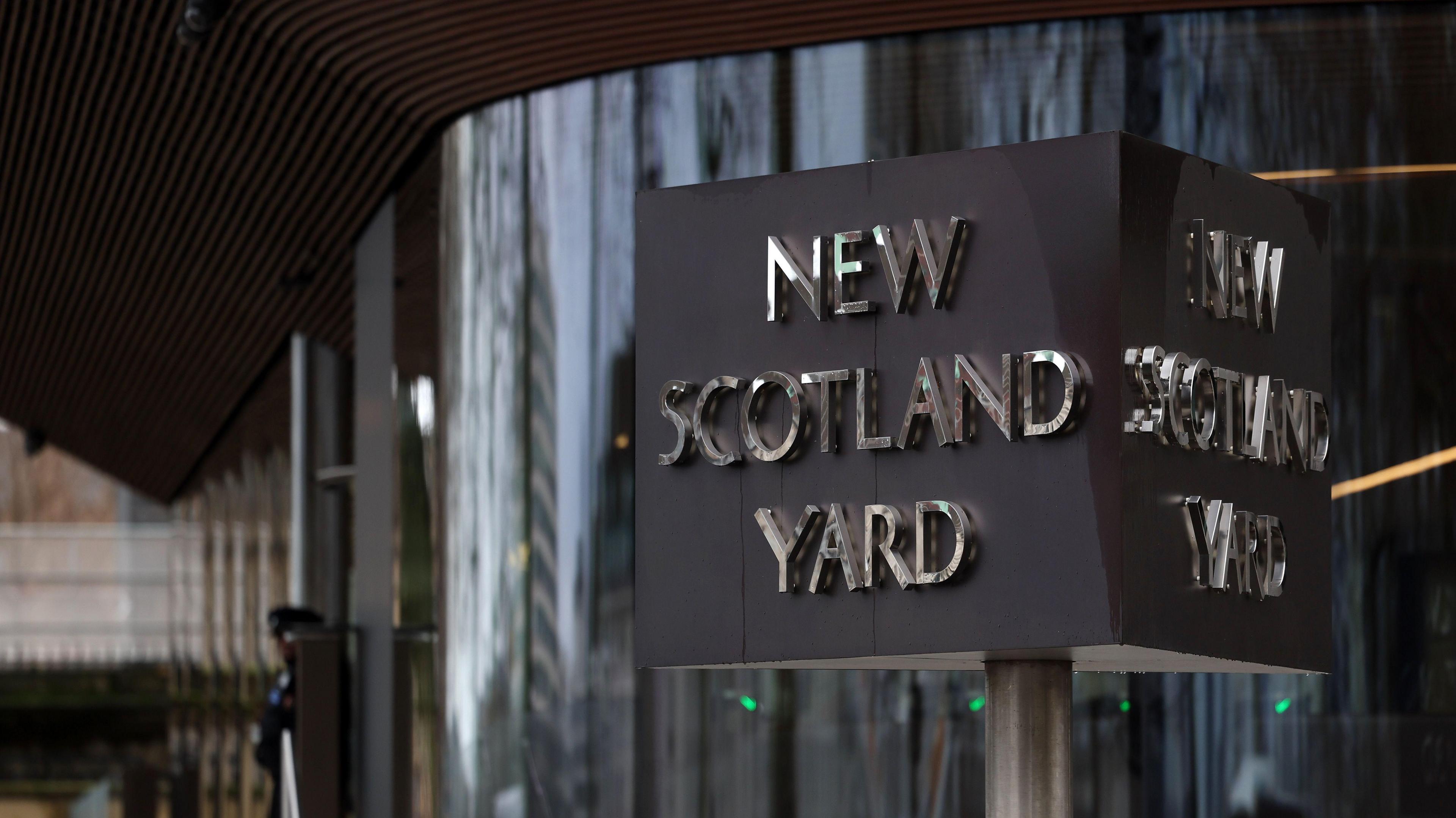 The New Scotland Yard sign outside the Metropolitan Police headquarters in London, with a police officer in the background.