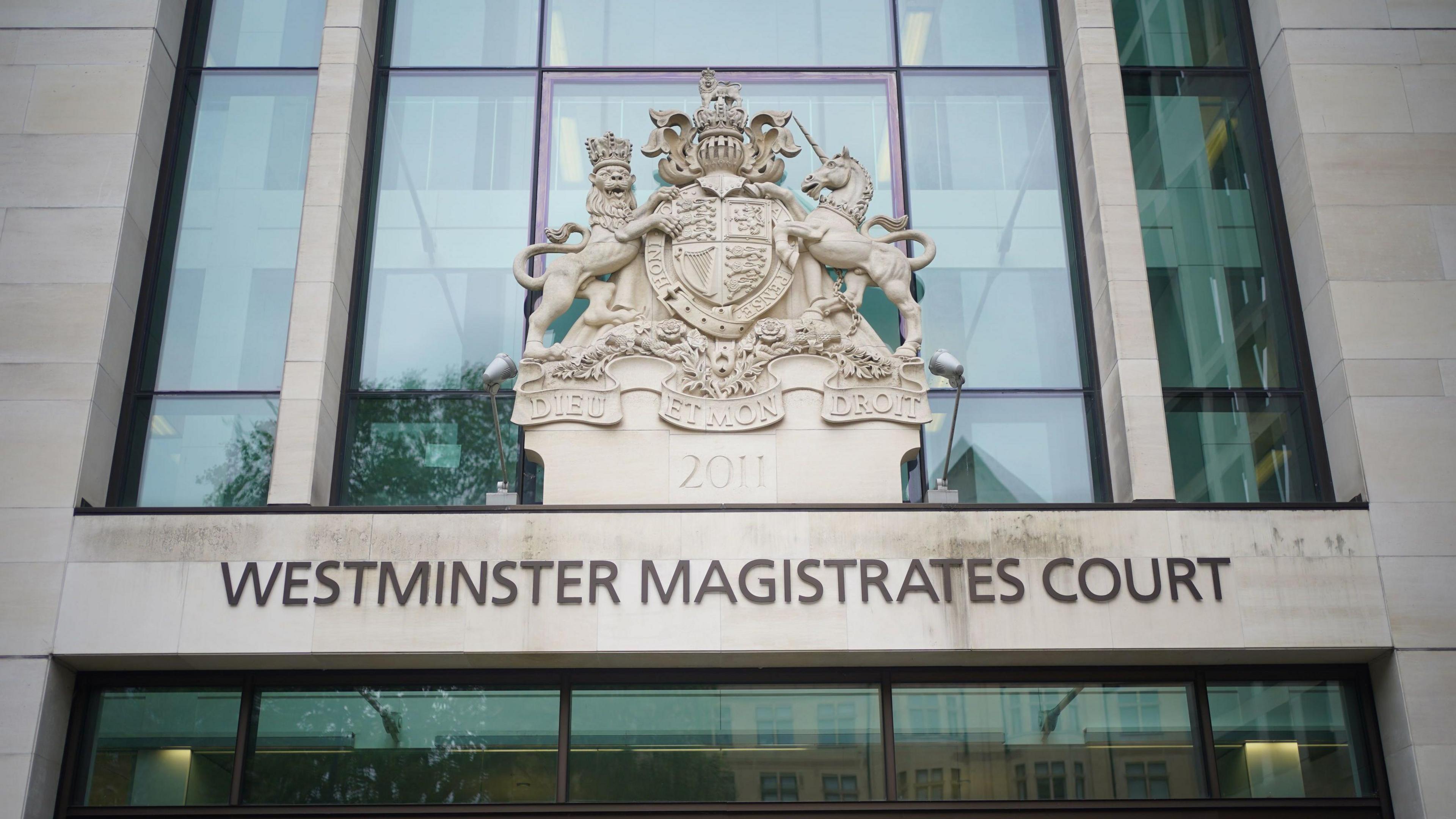 The exterior of Westminster Magistrates Court, with its name written above the entrance. Above the text, there's a large decorative coat of arms mounted on the building. The coat of arms shows a shield supported by a crowned lion on the left and a unicorn on the right, topped with a crown and a banner below that reads "DIEU ET MON DROIT."