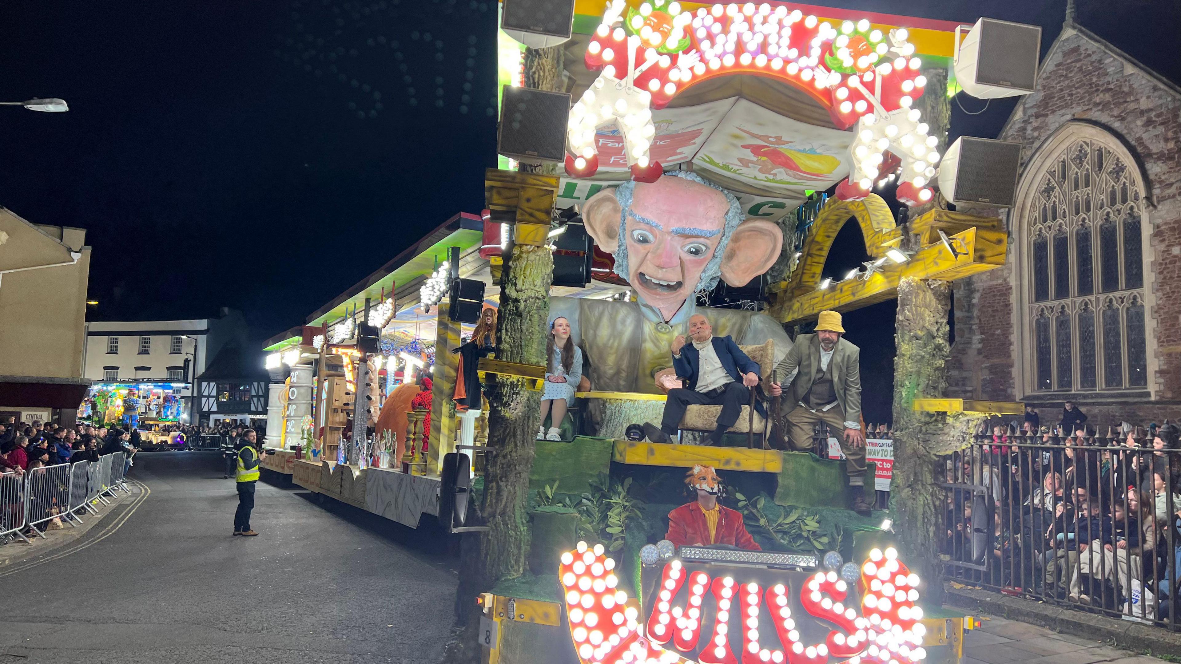 People ride on a float, which bears a large, crafted old man's head, as it drives down a street in Bridgwater and onlookers watch from behind barricades