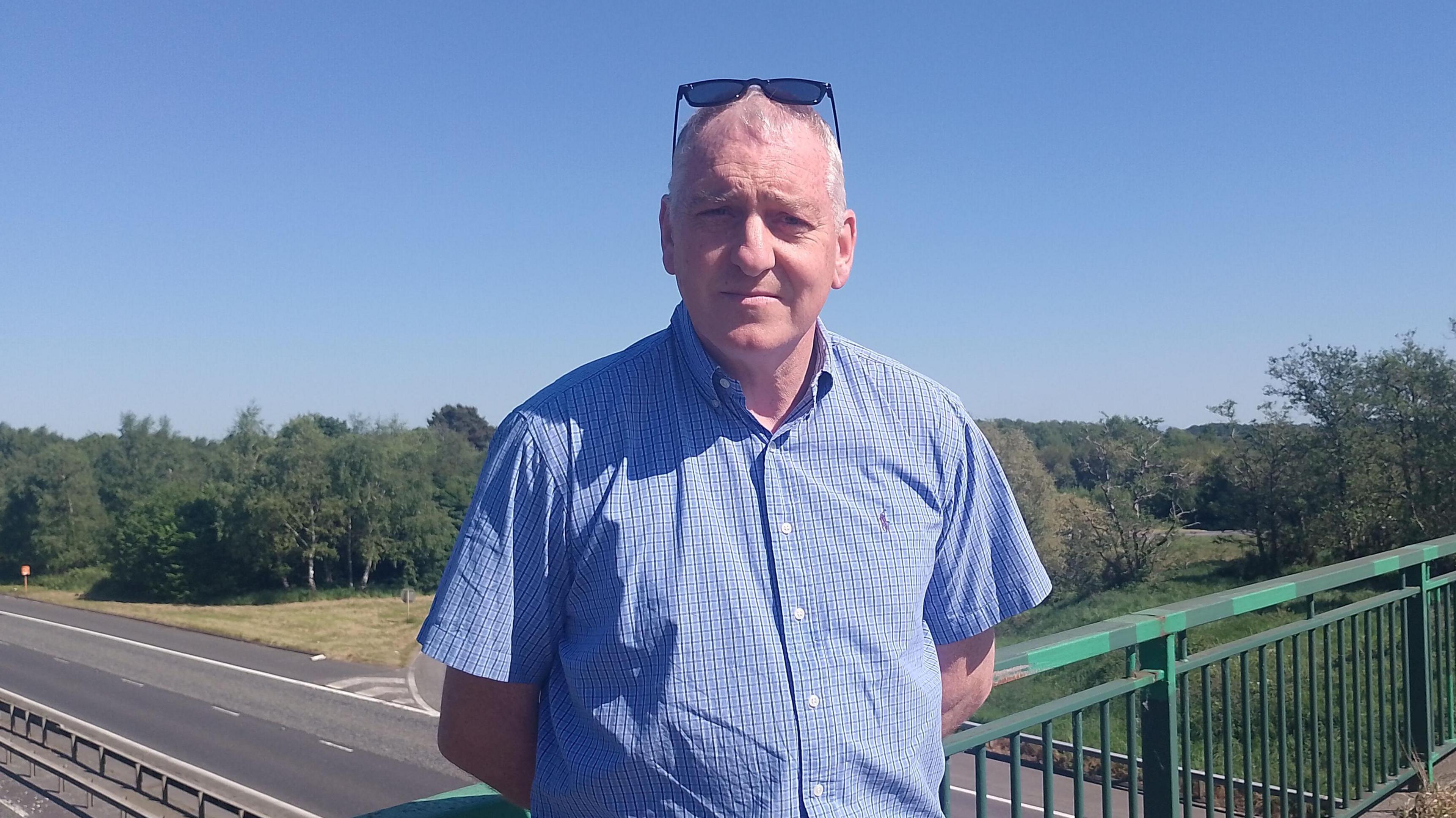 Lucas Vogan. He has short white hair, wearing sunglasses on his head and a light blue short sleeve shirt. He is standing on a motorway bridge with his hands behind his back.