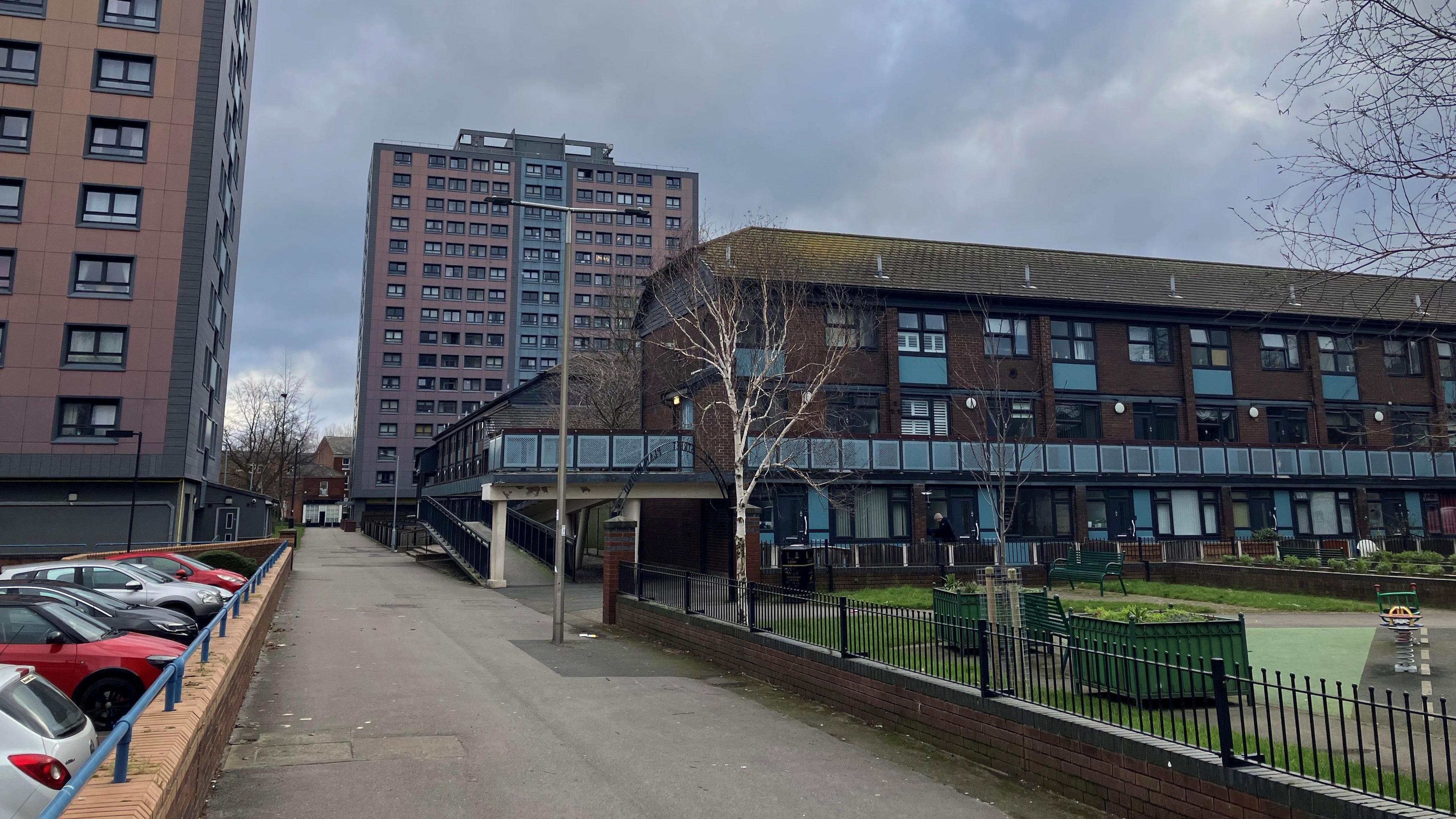 Photograph of homes on the Mottram Street estate in Stockport. The housing is a three-storey brown brick building with a balcony running around the second storey which you can access via a ramp. At the front there is a green area with children's play equipment and planters. 