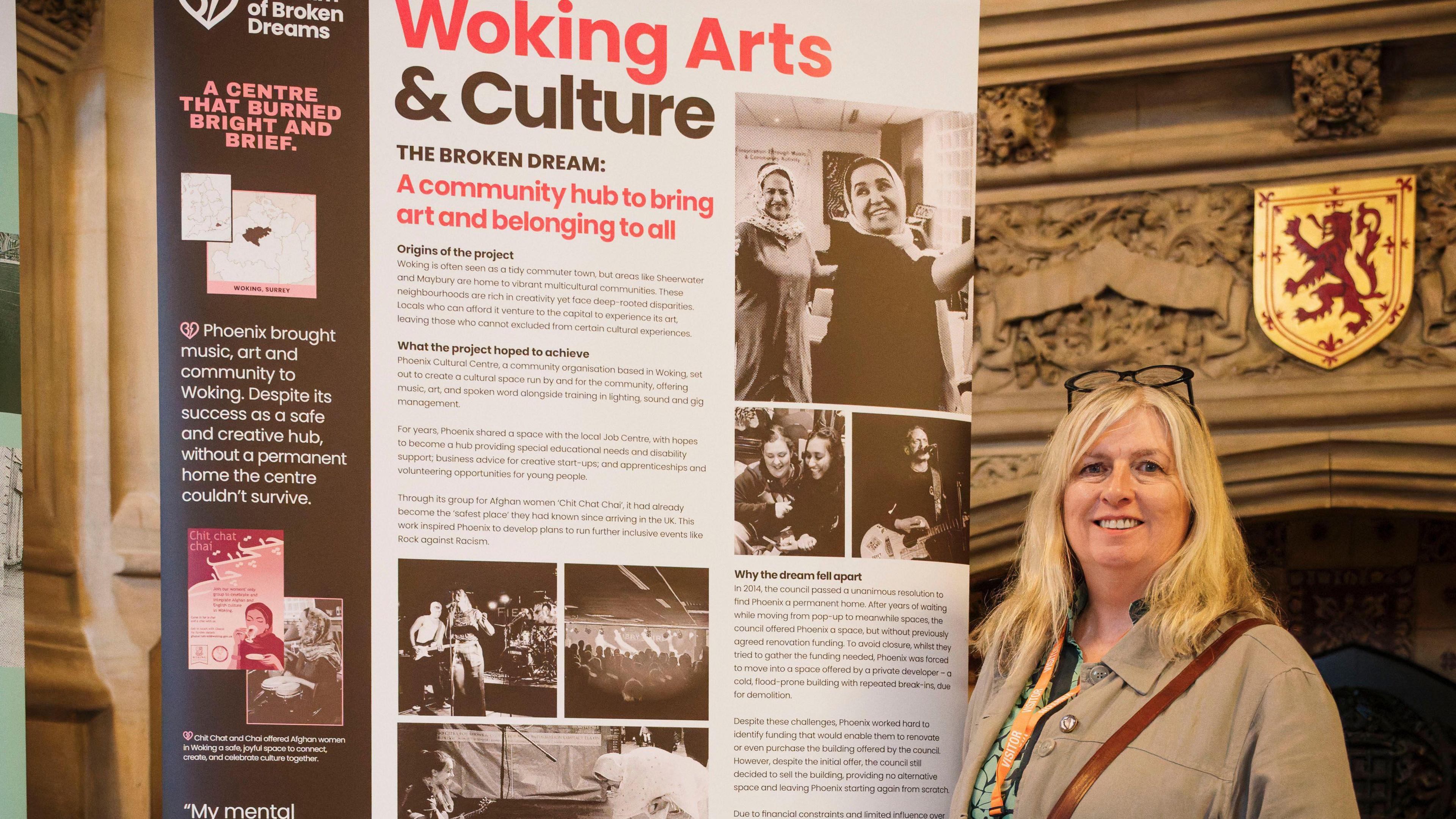 A blonde woman is standing next to a large red, white and black poster board titled "Woking Arts & Culture".