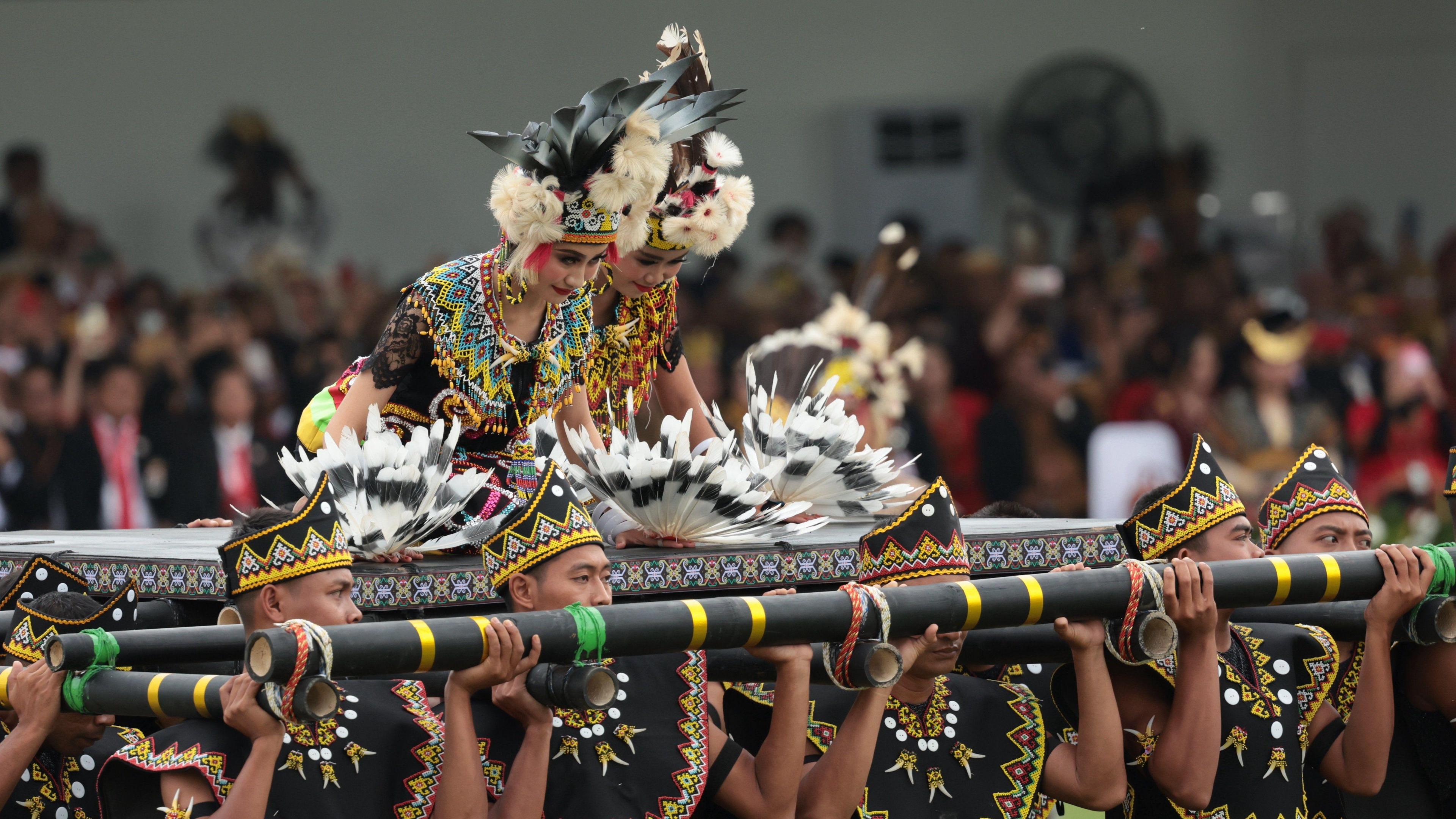 Two dancers in costume sit upon a platform carried by men. 