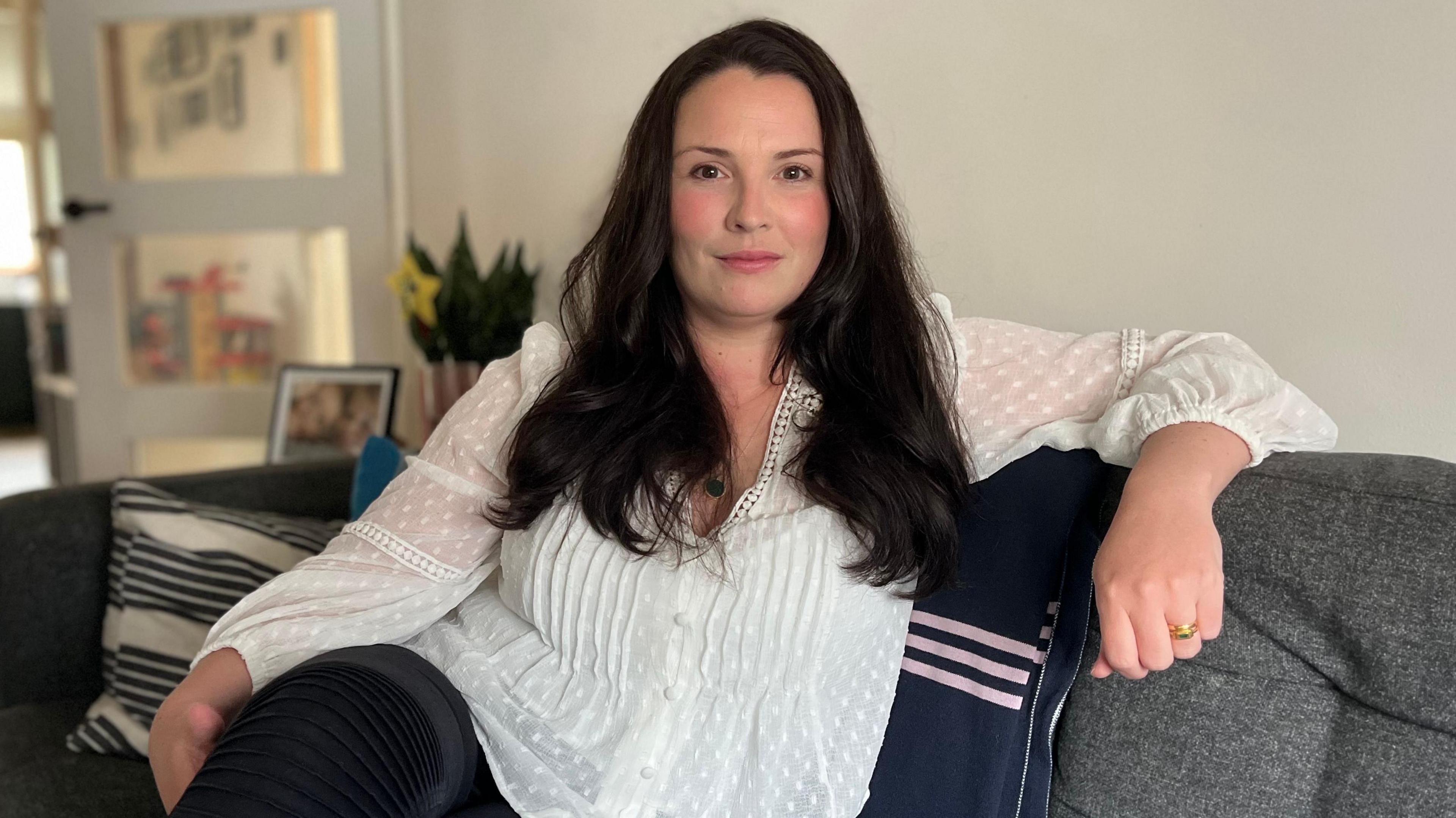 The image shows a woman with long dark brown hair laying on a sofa, her left arm resting on the back of it. She is wearing a white blouse with see-through sleeves and her trousers are black. A golden ring can also be seen on her left hand's ring finger. The couch she is sitting on is grey and a black and white striped cushion and the corner of a deep blue one can be seen behind her. In the background, a daffodil in a pot can be seen, as well as a blurry picture frame.